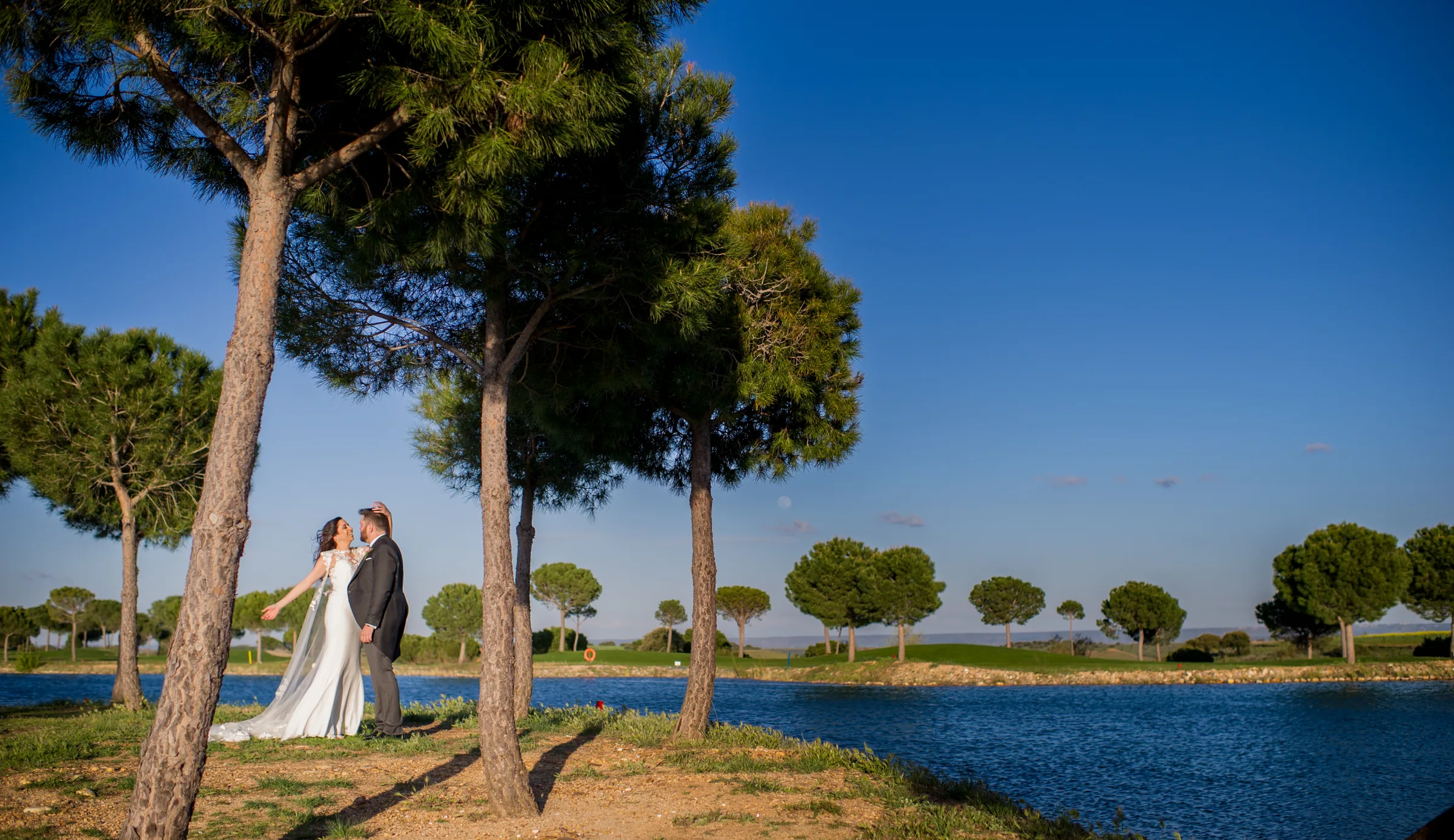 Los novios junto al lago bajo los pinos con cielo azul en Retamares Experience.