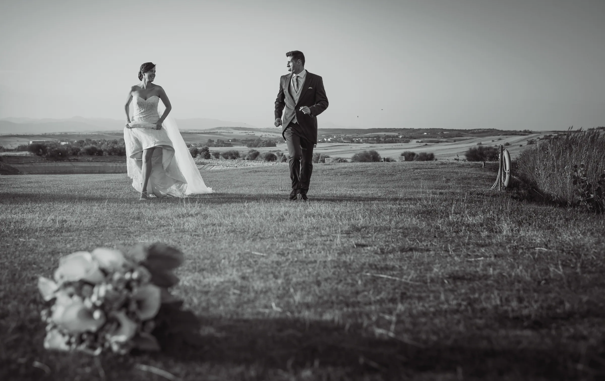 Los novios caminan por el campo en blanco y negro con el ramo en primer plano en Retamares Experience.