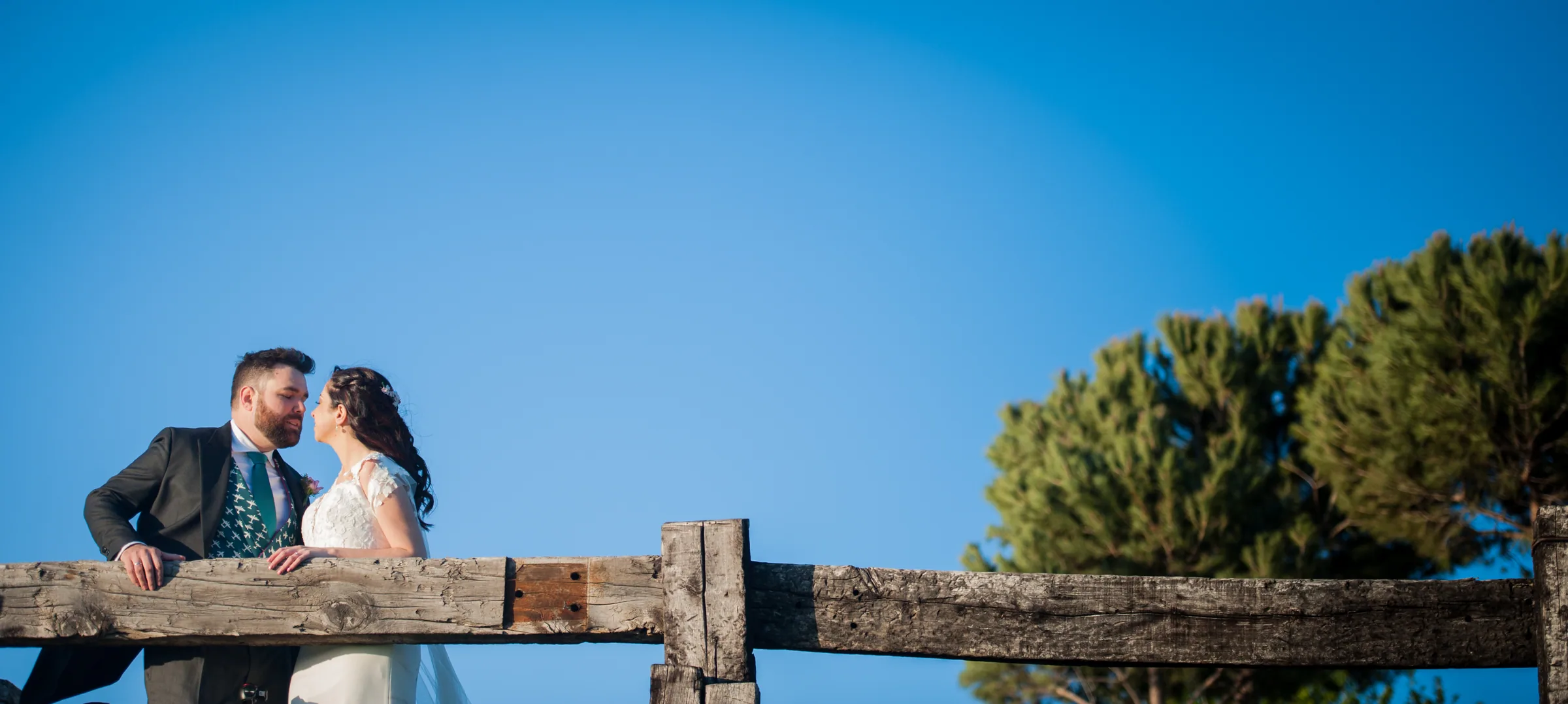 Los novios se besan tras una valla de madera con pinos y cielo azul en Retamares Experience.