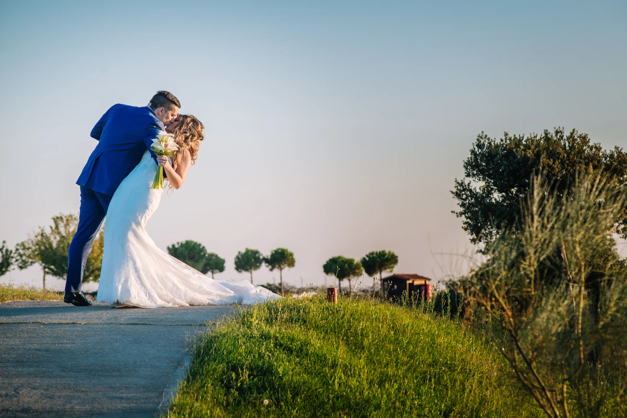 Los novios se besan inclinados sobre un camino con cielo azul y árboles en Retamares Experience.