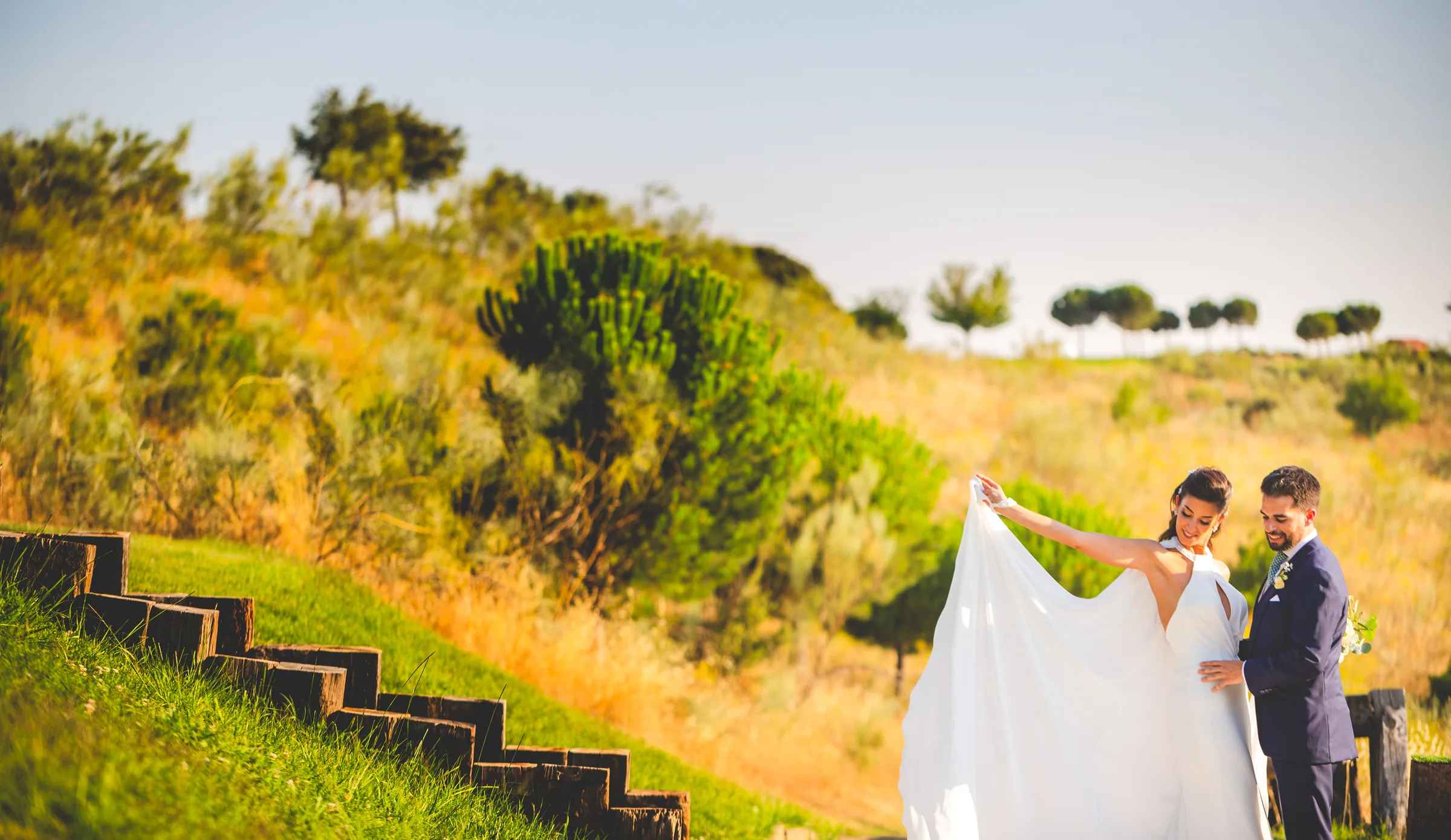 Los novios en una ladera de hierba dorada con la novia sujetando el velo en Retamares Experience.