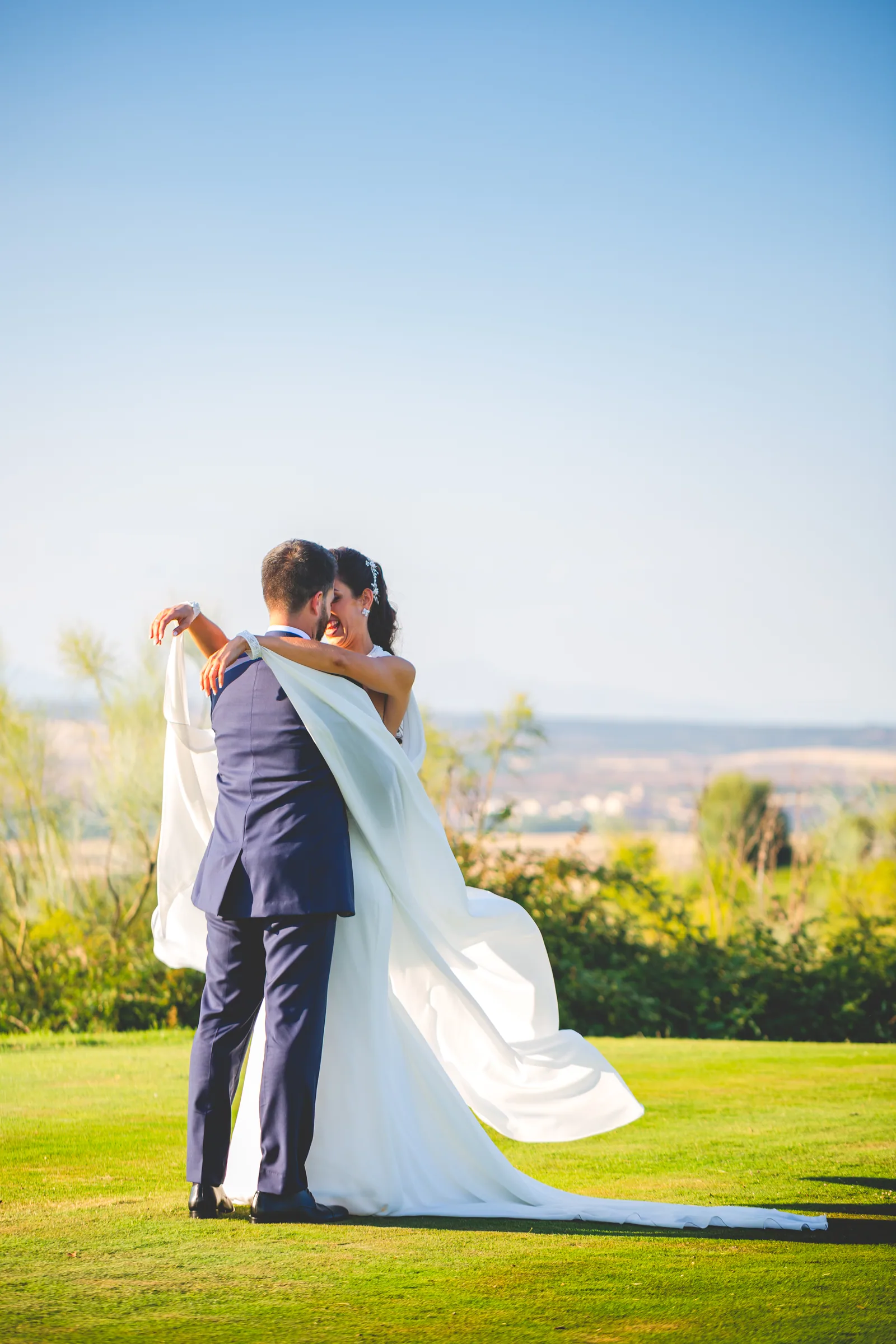 Los novios abrazados sobre el césped con el velo y el cielo azul en Retamares Experience.