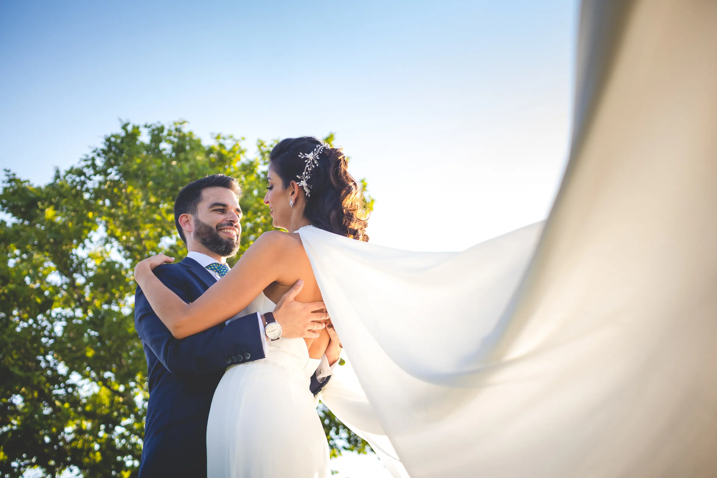 Los novios se abrazan bajo el velo ondeando con las copas de los árboles detrás en Retamares Experience.