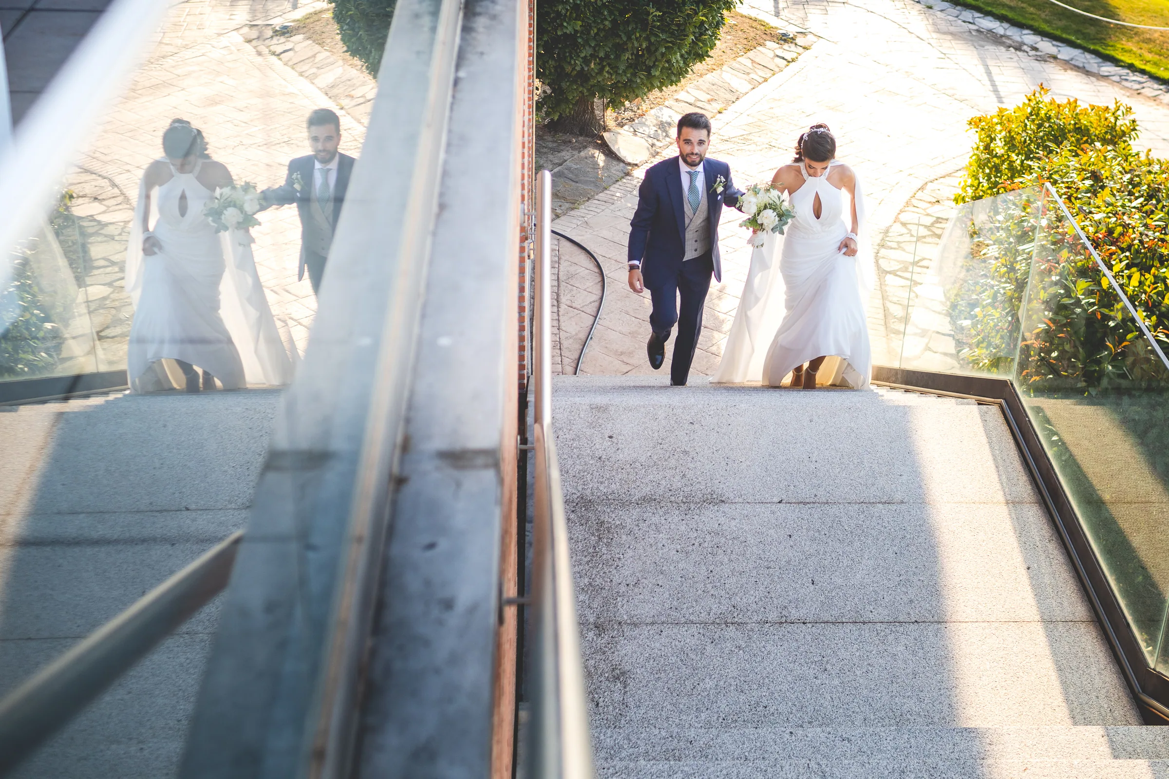 Los novios suben por una escalera exterior vistos desde arriba, con su reflejo en el cristal en Retamares Experience.