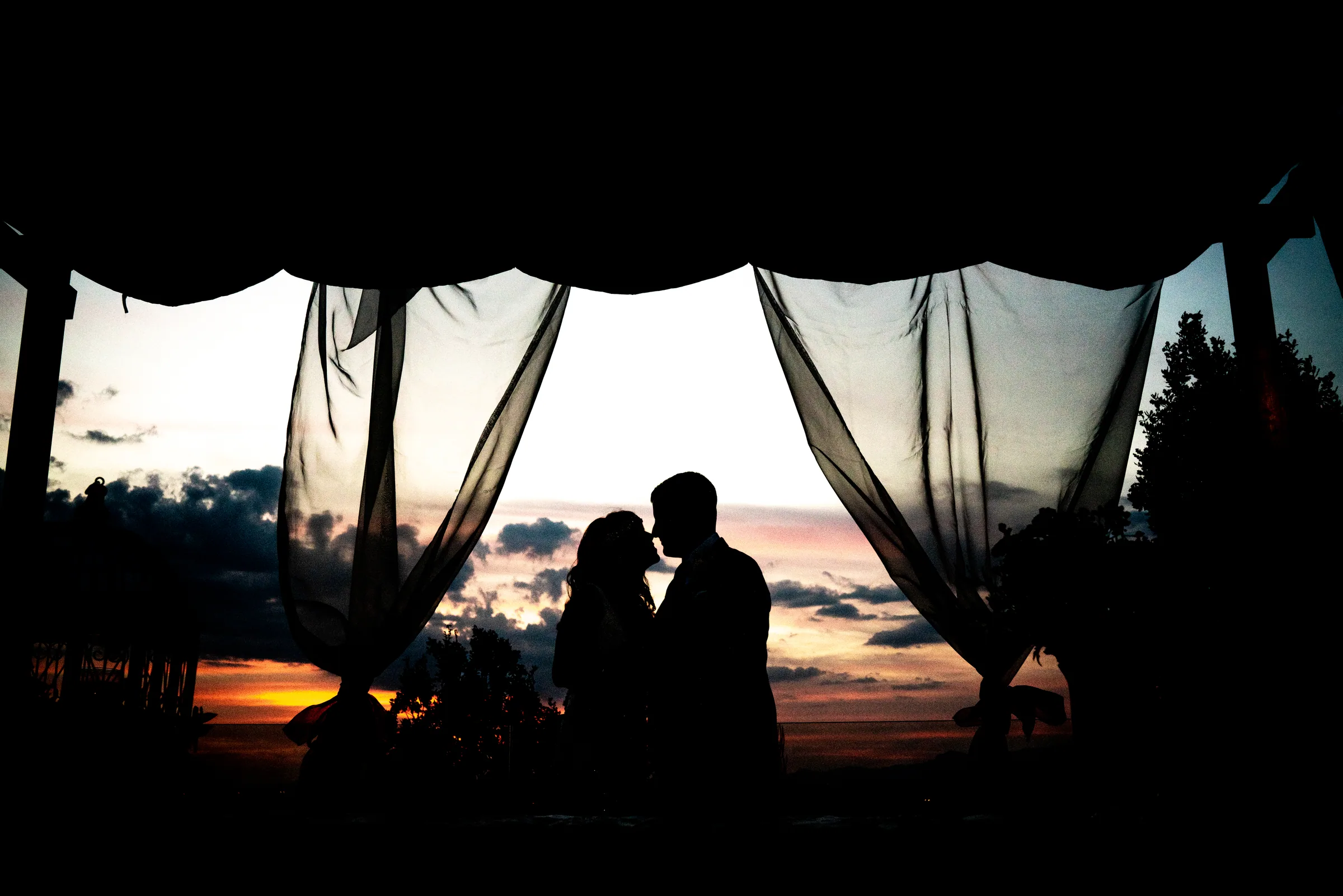 Silueta de los novios besándose entre cortinas de gasa con el atardecer detrás en Retamares Experience.