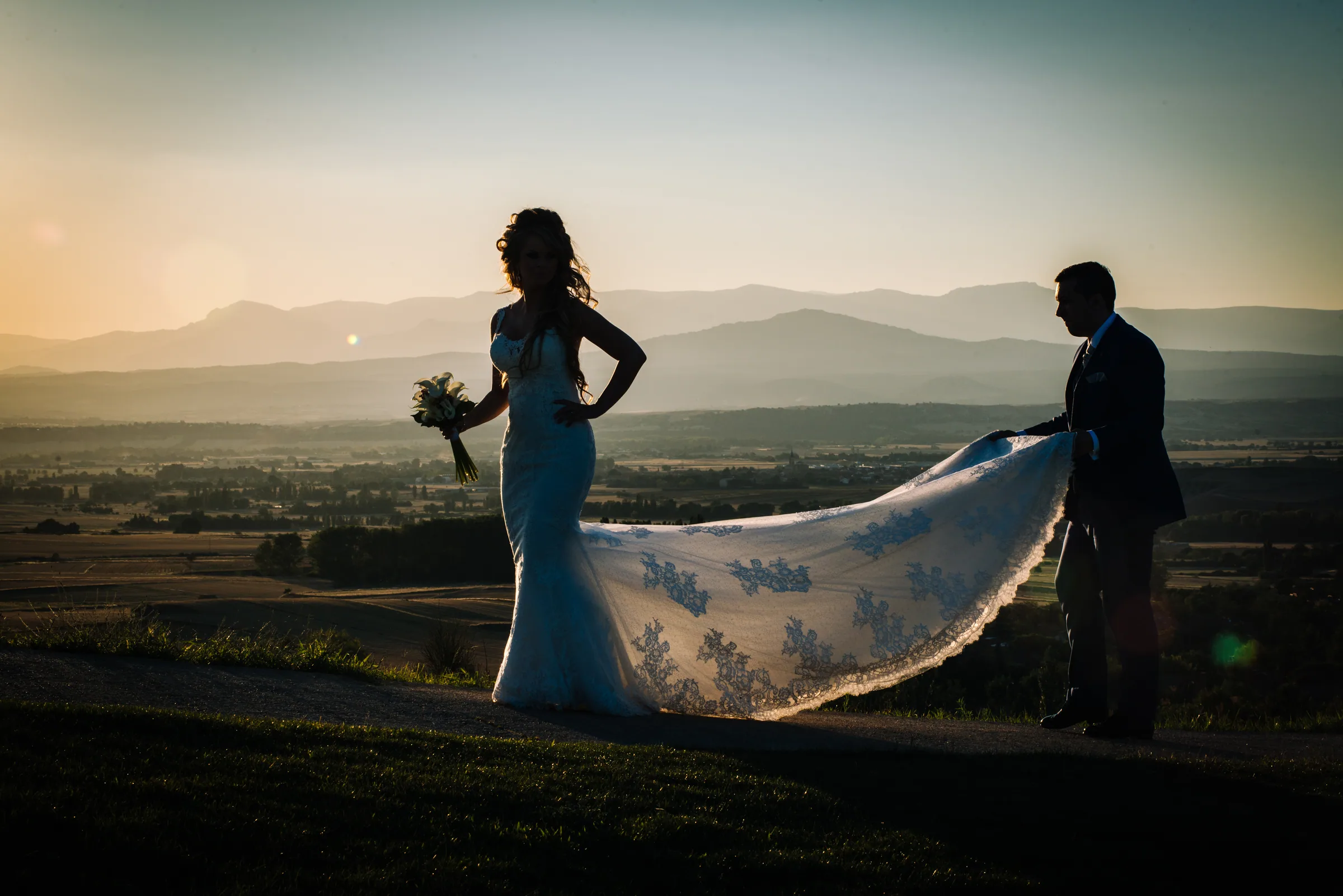Los novios a contraluz con la cola del vestido extendida y la sierra al fondo en Retamares Experience.