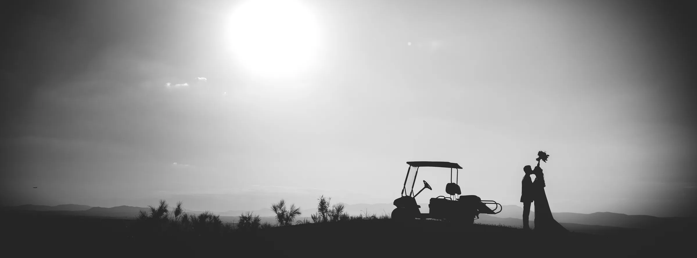 Panorámica en blanco y negro del sol sobre el campo con un buggy de golf y la silueta de los novios en Retamares Experience.