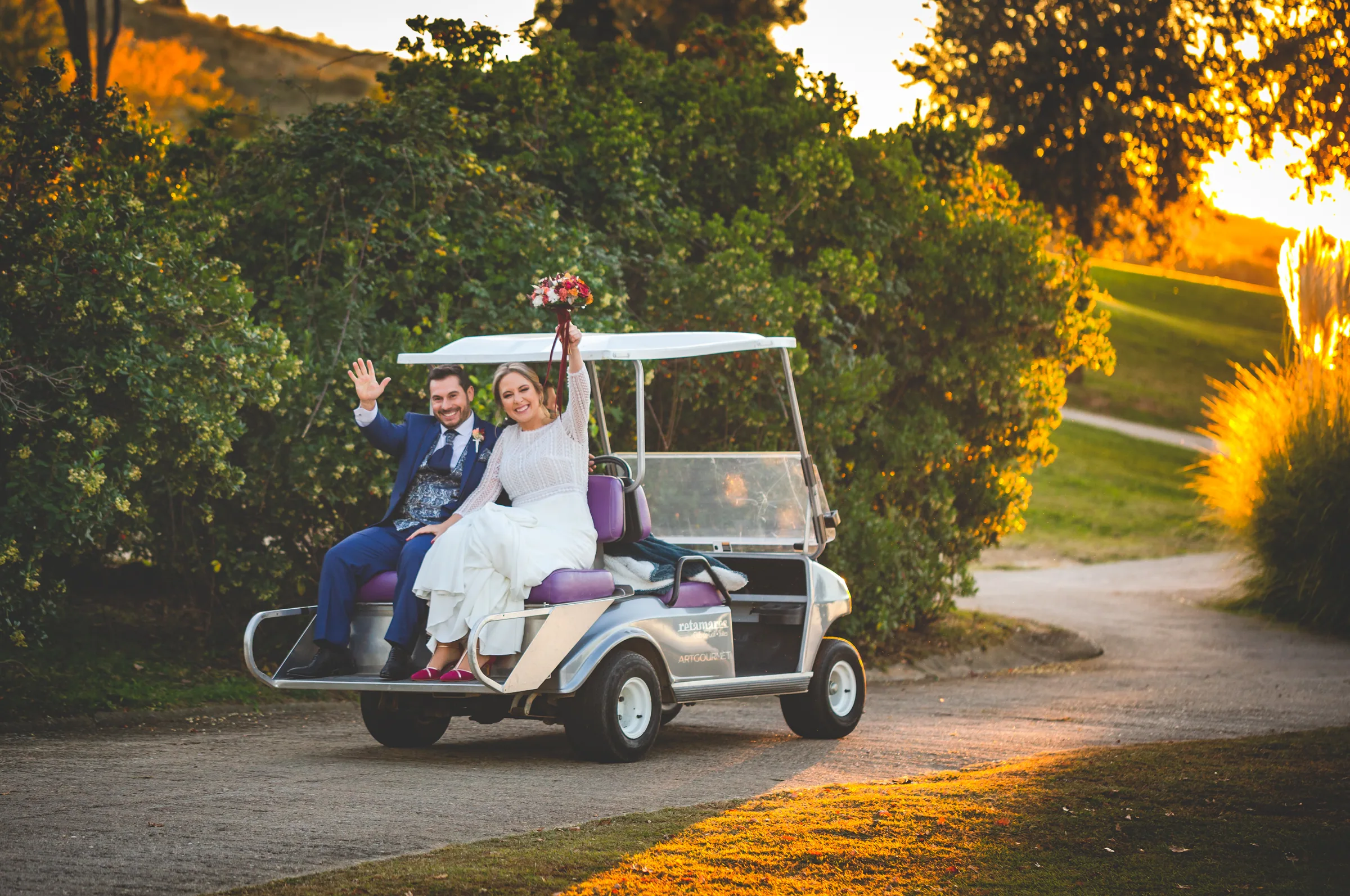 Los novios saludan desde un buggy de golf por un camino entre vegetación en Retamares Experience.
