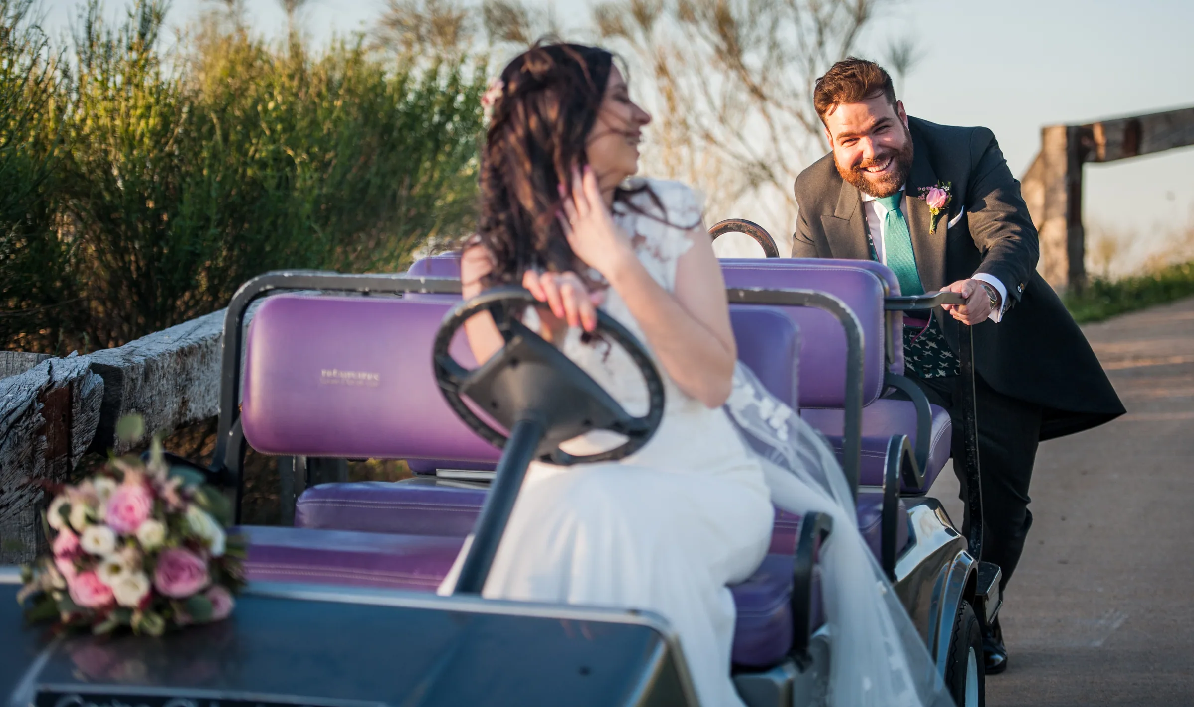 La novia sentada en un buggy de golf con su ramo mientras el novio sonríe a su lado en Retamares Experience.