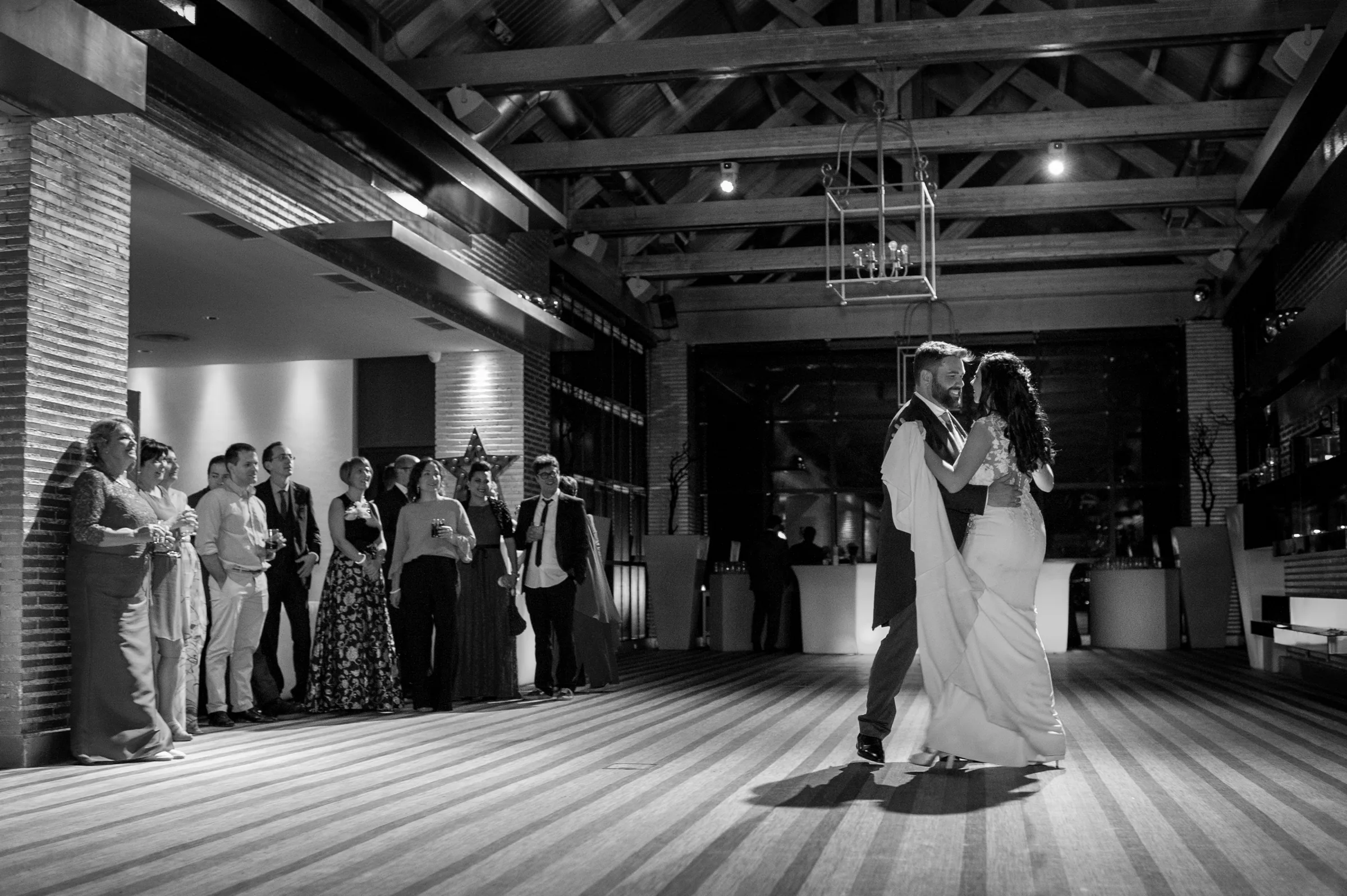 Primer baile en blanco y negro en un salón con vigas de madera y los invitados alrededor en Retamares Experience.