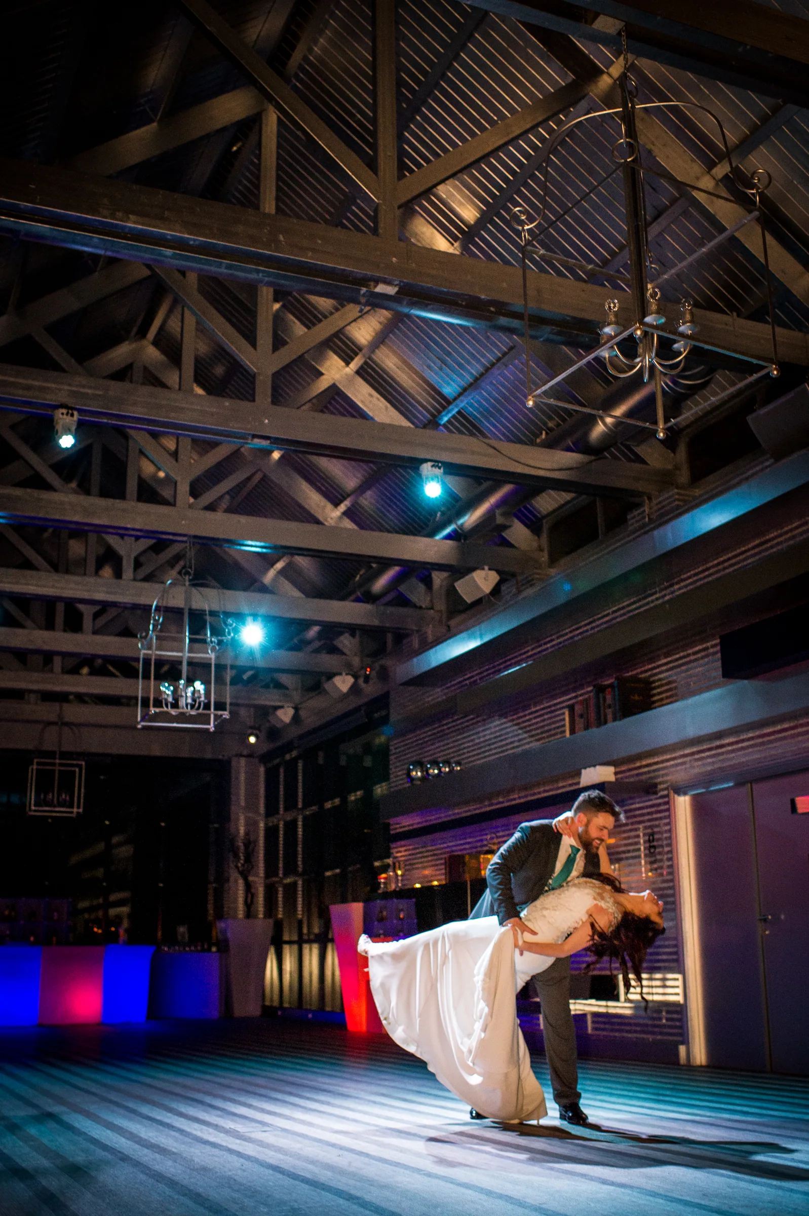 El novio inclina a la novia en la pista de baile bajo las vigas del salón con luces de colores en Retamares Experience.