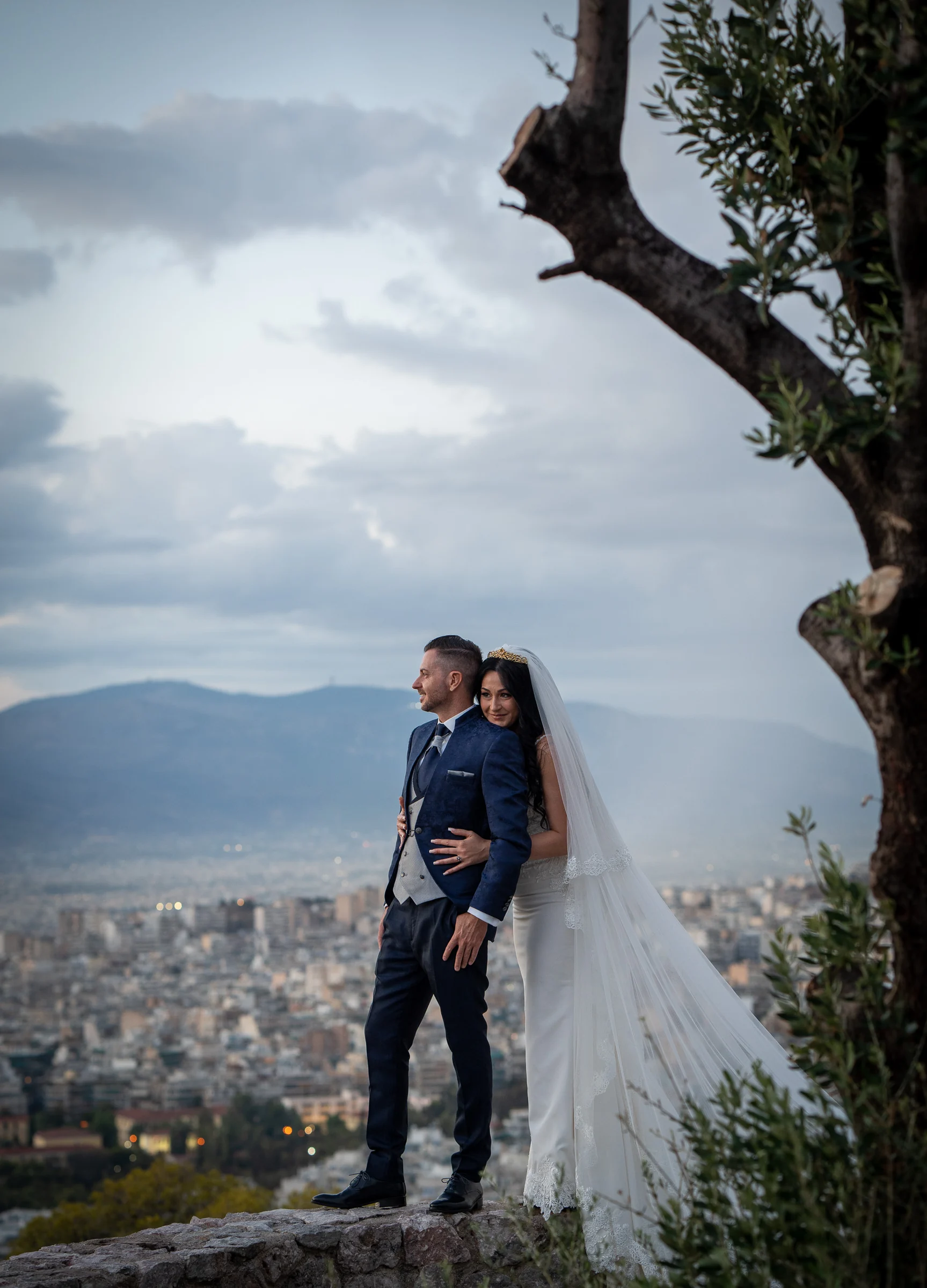 Sara abraza a Edu junto a un olivo mientras contemplan el panorama de Atenas durante su postboda en Grecia.