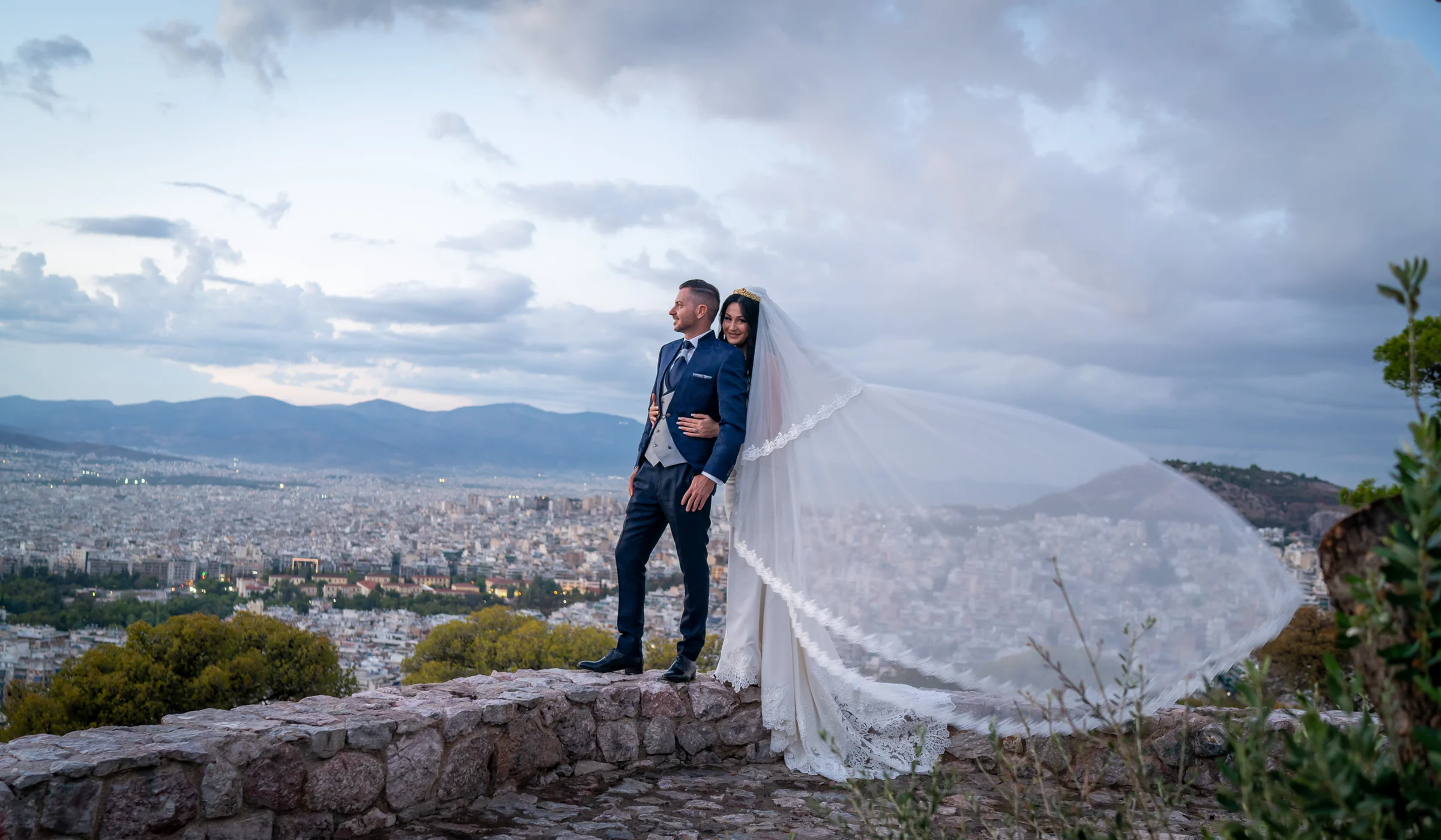 Sara y Edu posan en un mirador de Atenas mientras el velo de Sara se extiende con el viento sobre la panorámica de la ciudad.