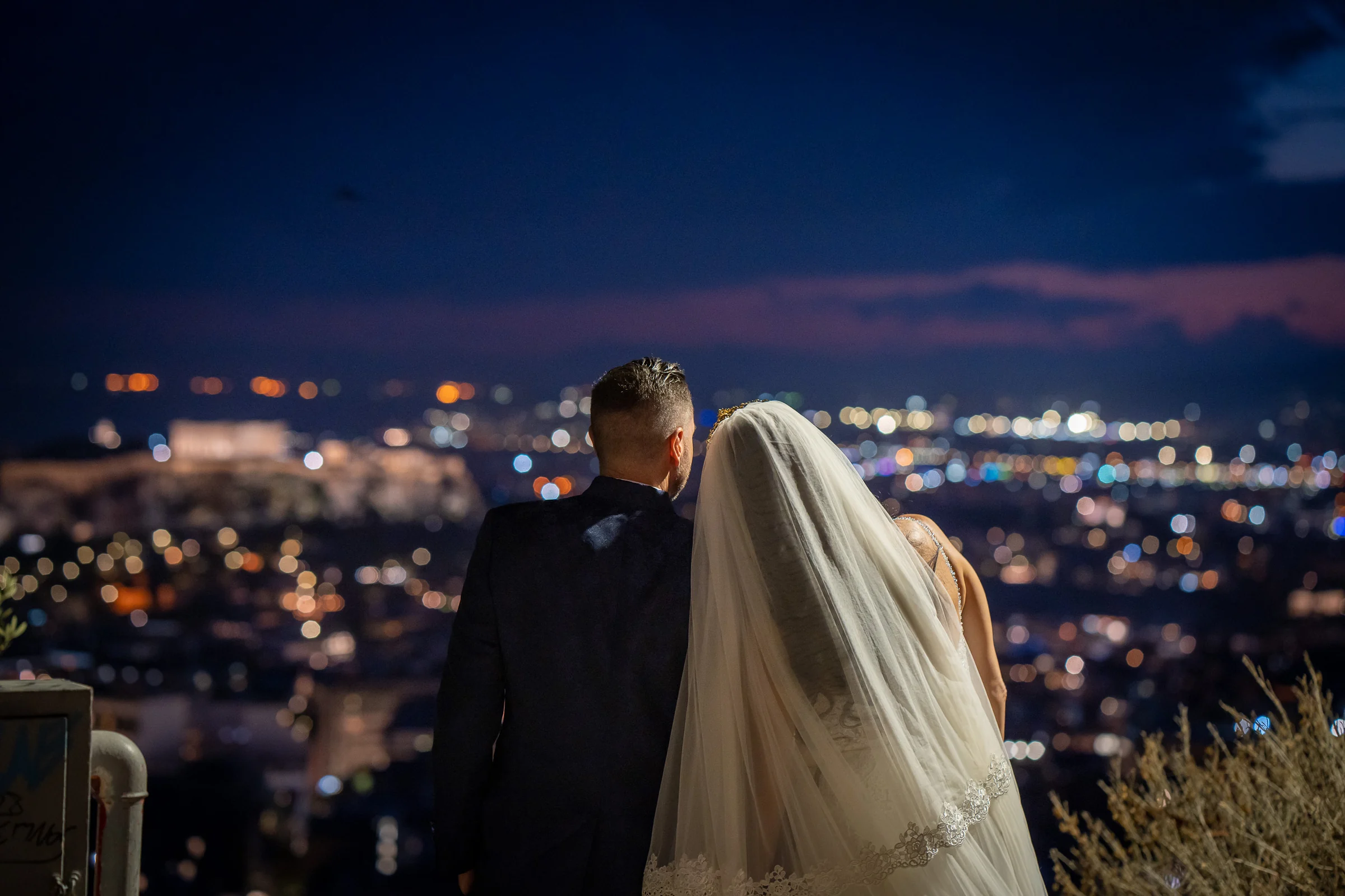 Sara y Edu contemplan de espaldas las luces nocturnas de Atenas durante su postboda en Grecia.