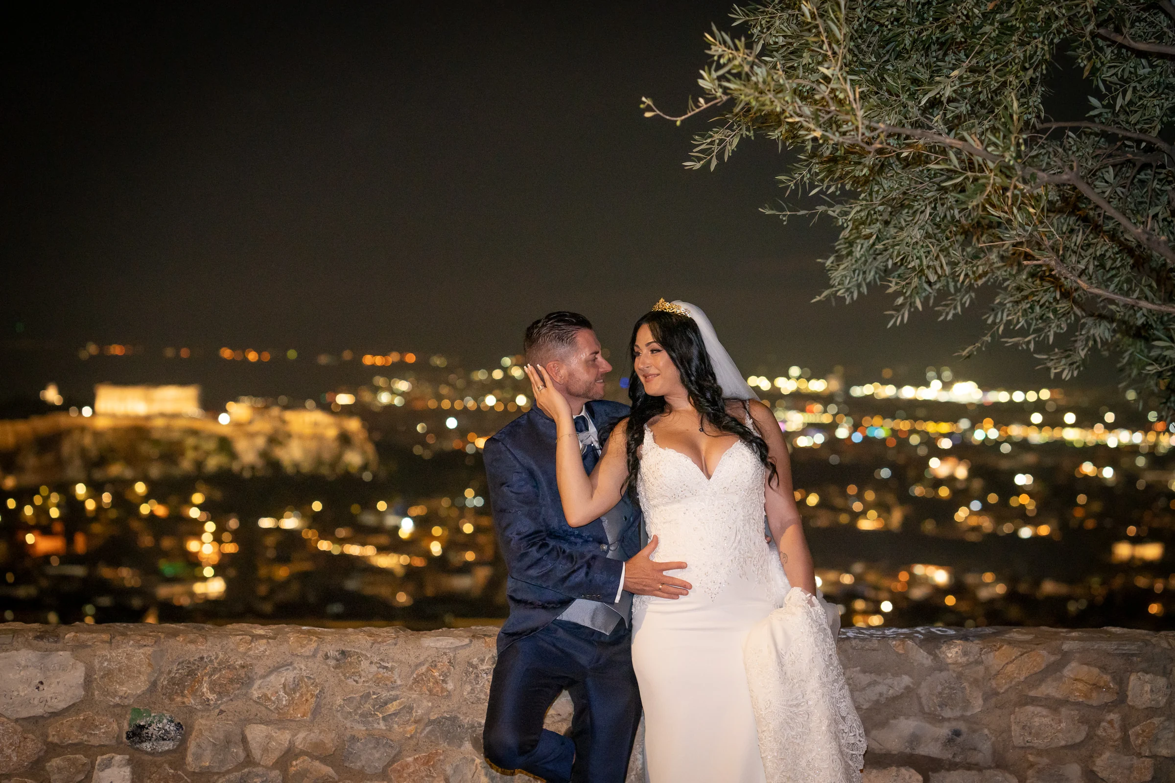 Sara acaricia el rostro de Edu en un retrato nocturno con las luces de Atenas y la Acrópolis al fondo durante su postboda en Grecia.