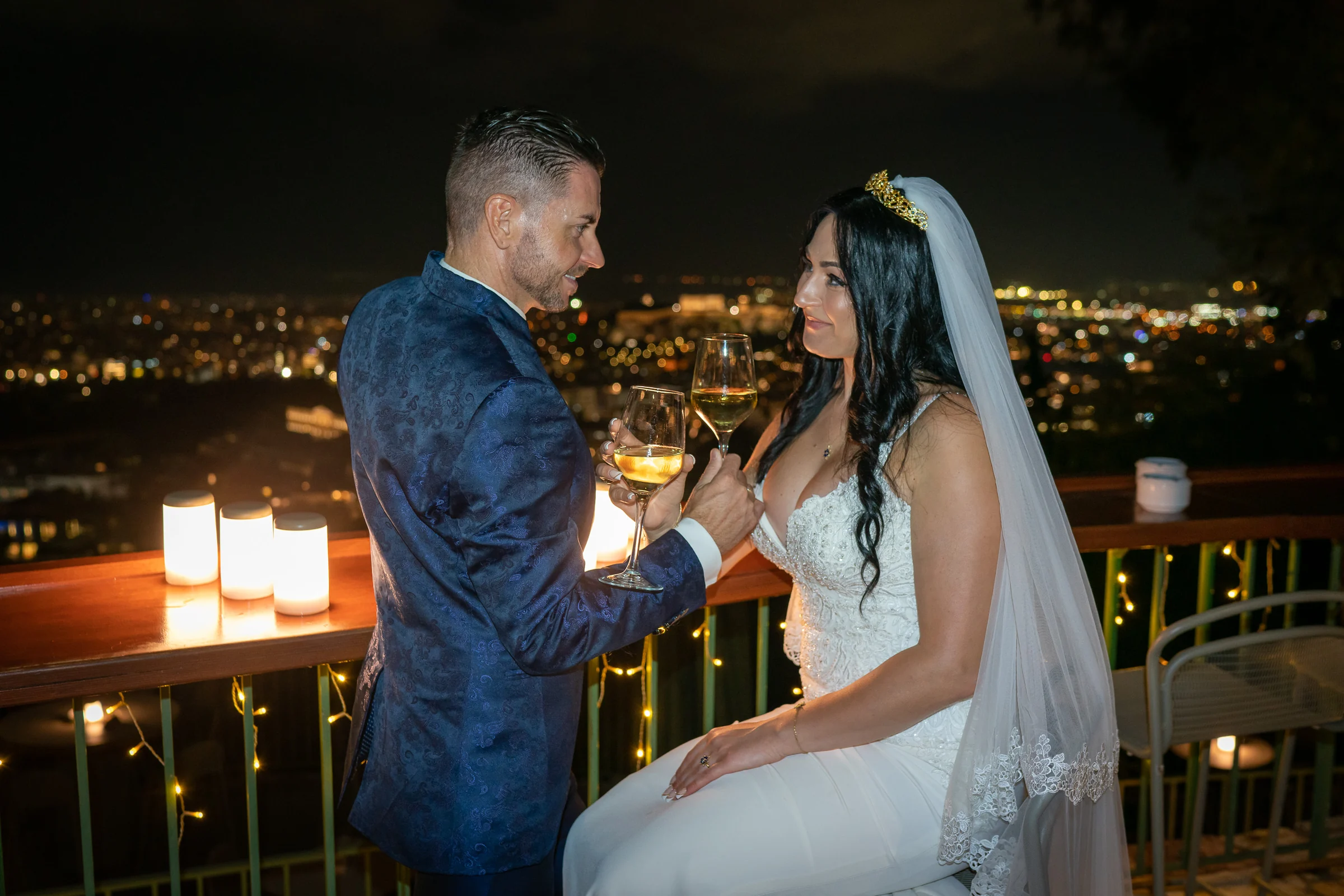 Sara y Edu brindan con vino en una terraza iluminada con Atenas de noche al fondo durante su postboda en Grecia.
