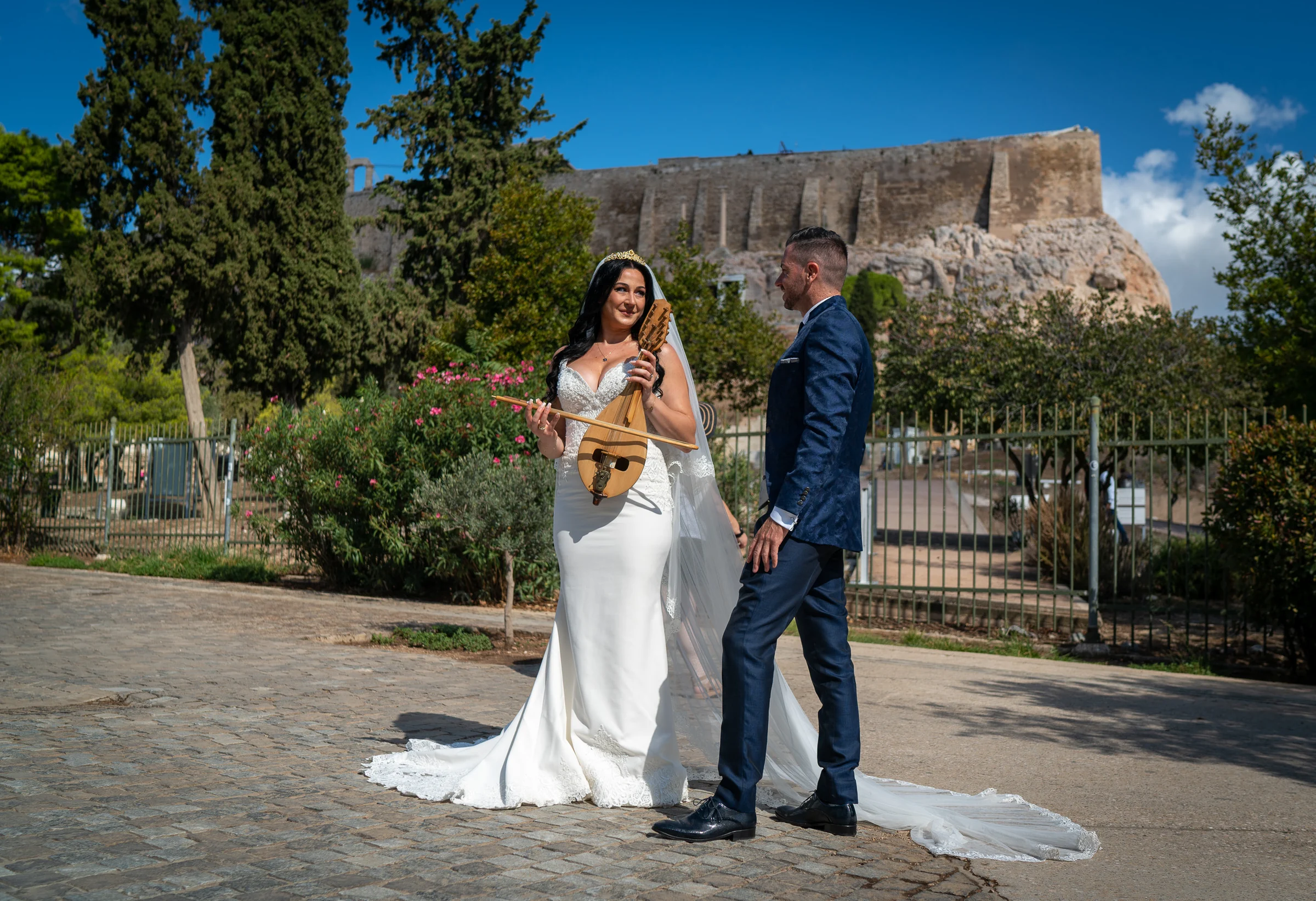 Sara sostiene un instrumento tradicional griego junto a Edu con la Acrópolis de Atenas al fondo durante su postboda en Grecia.