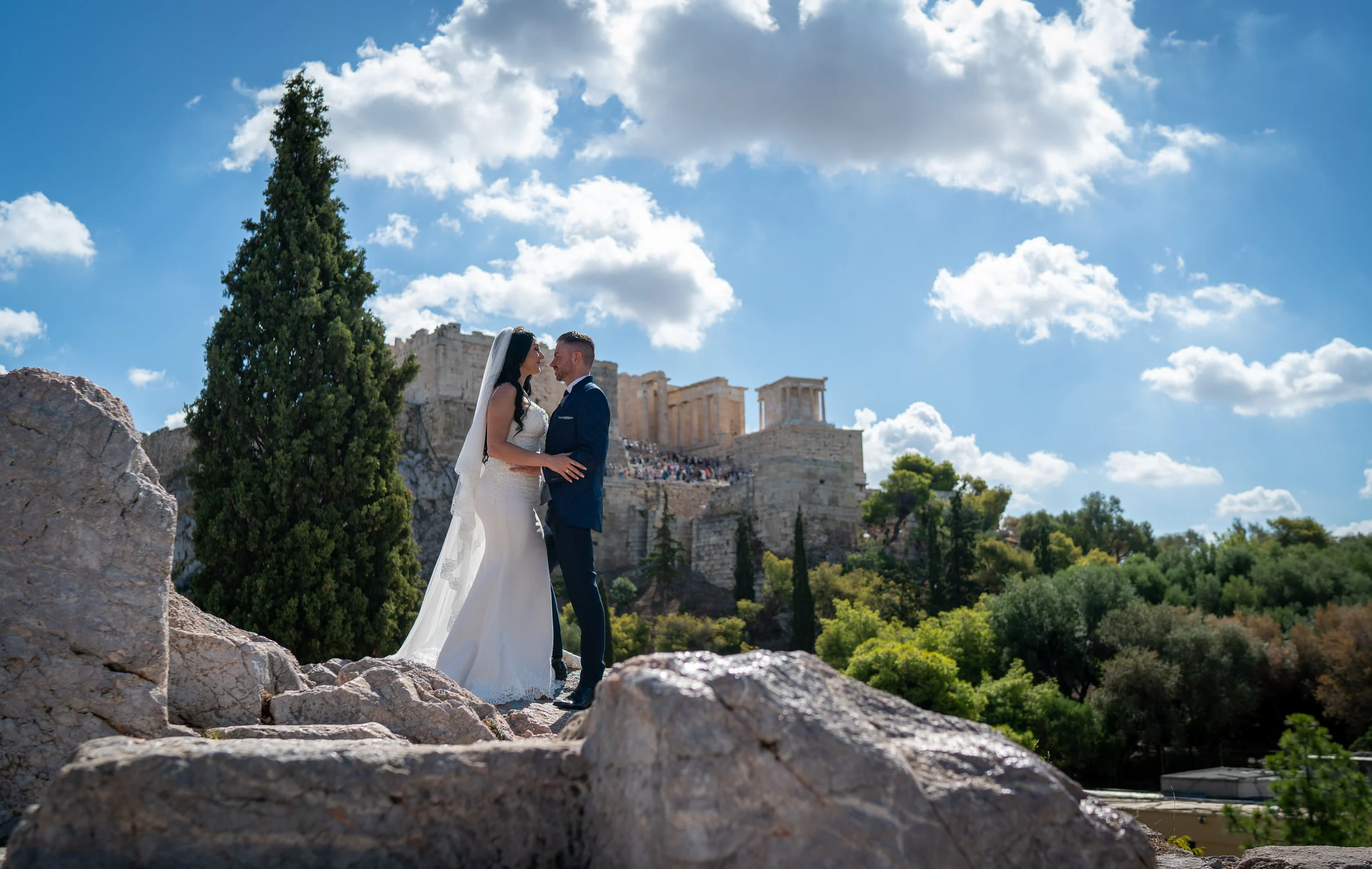 Sara y Edu se abrazan sobre las rocas con la Acrópolis de Atenas al fondo durante su postboda en Grecia.