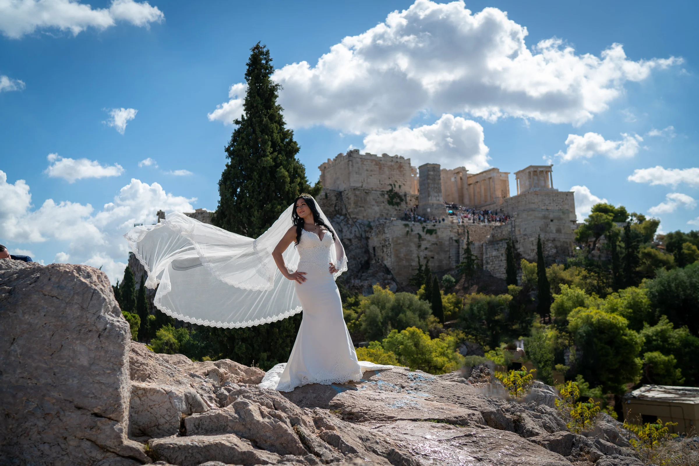 Sara posa sola con el velo extendido por el viento y la Acrópolis de Atenas al fondo durante la postboda en Grecia.