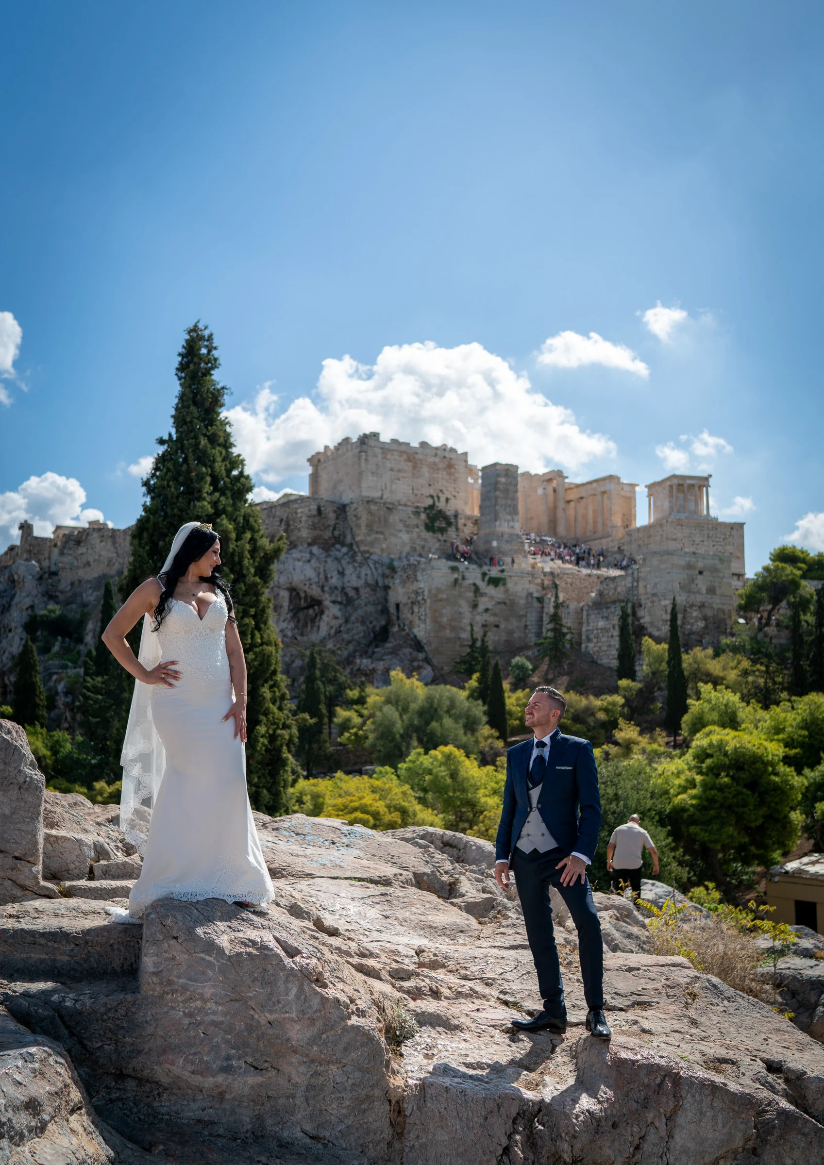 Sara y Edu se miran desde distintos niveles de una zona rocosa con la Acrópolis de Atenas al fondo durante su postboda.