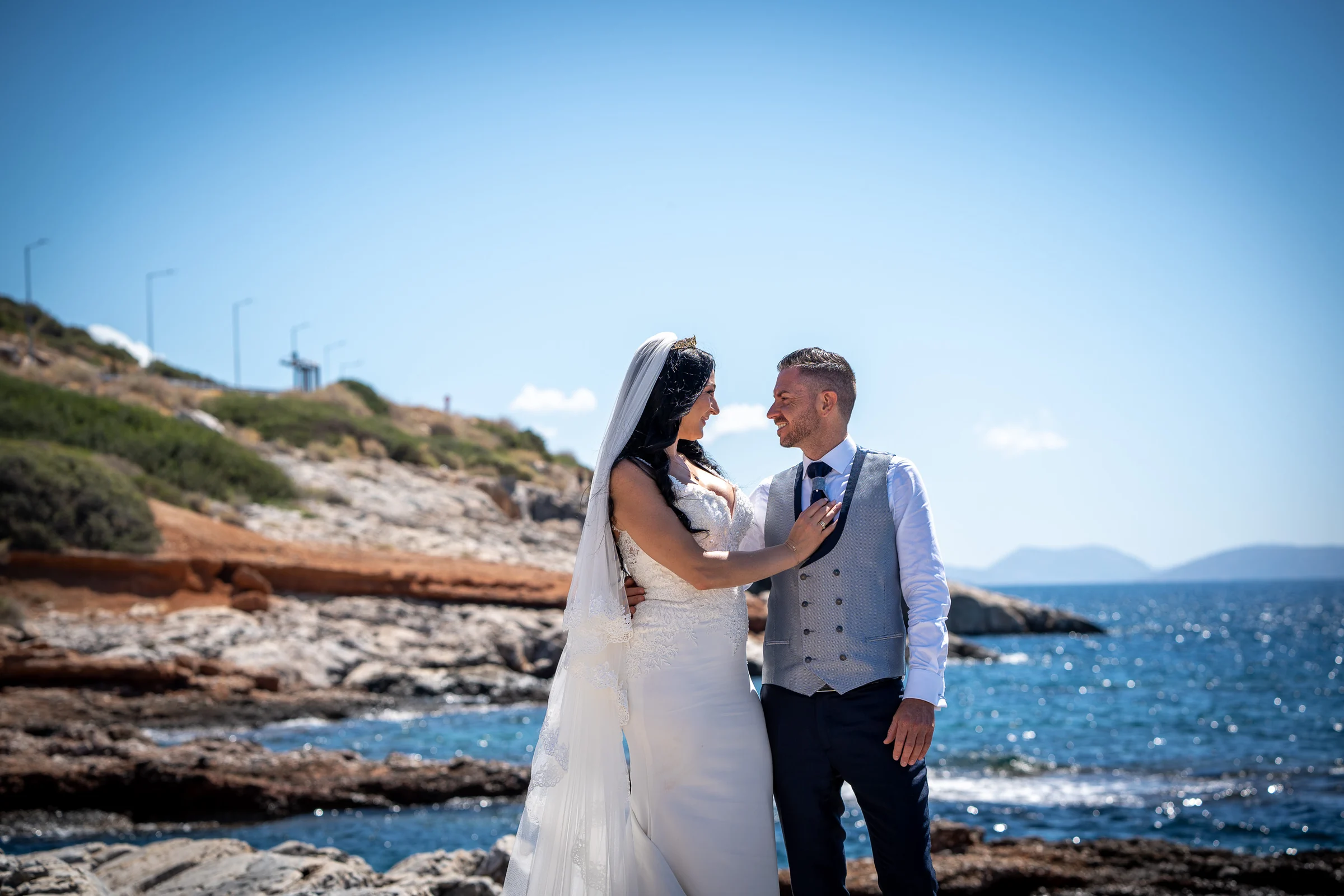 Sara y Edu se miran junto a una costa rocosa con el mar Egeo al fondo durante su postboda en Grecia.