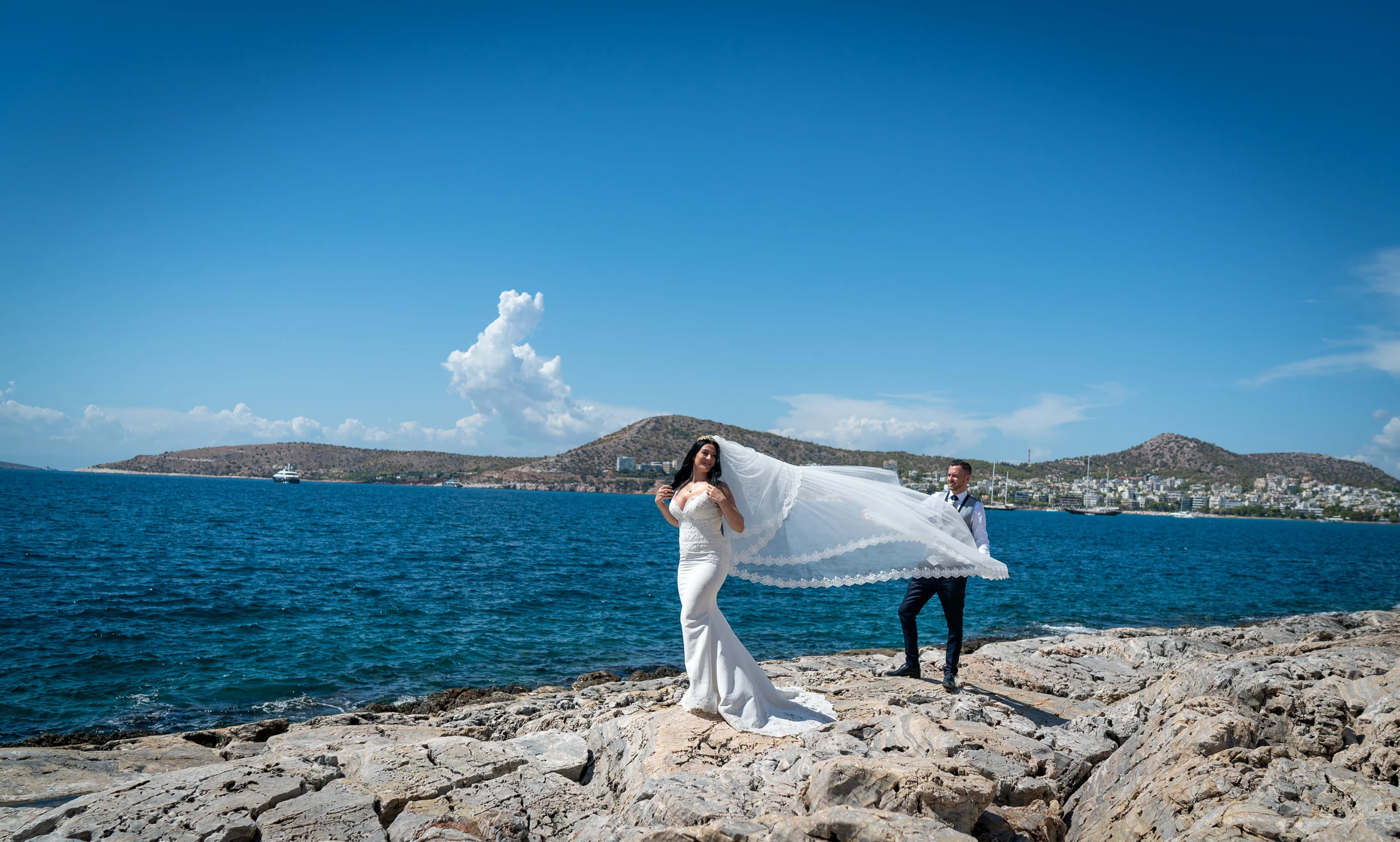 Sara posa sobre las rocas mientras Edu sostiene su velo al viento frente al mar Egeo durante su postboda en Grecia.