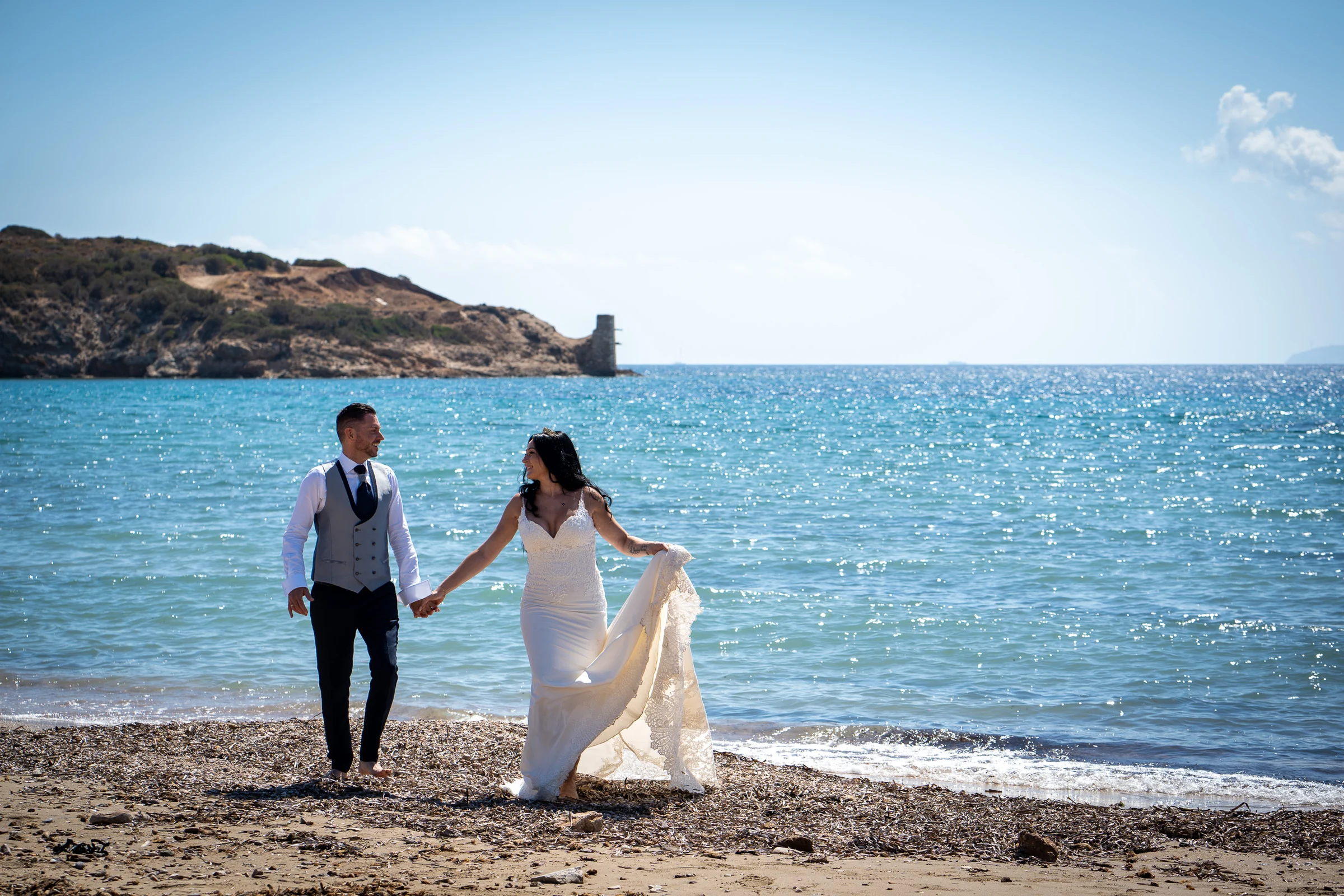 Sara y Edu caminan de la mano por una playa junto al mar Egeo durante su postboda en Grecia.