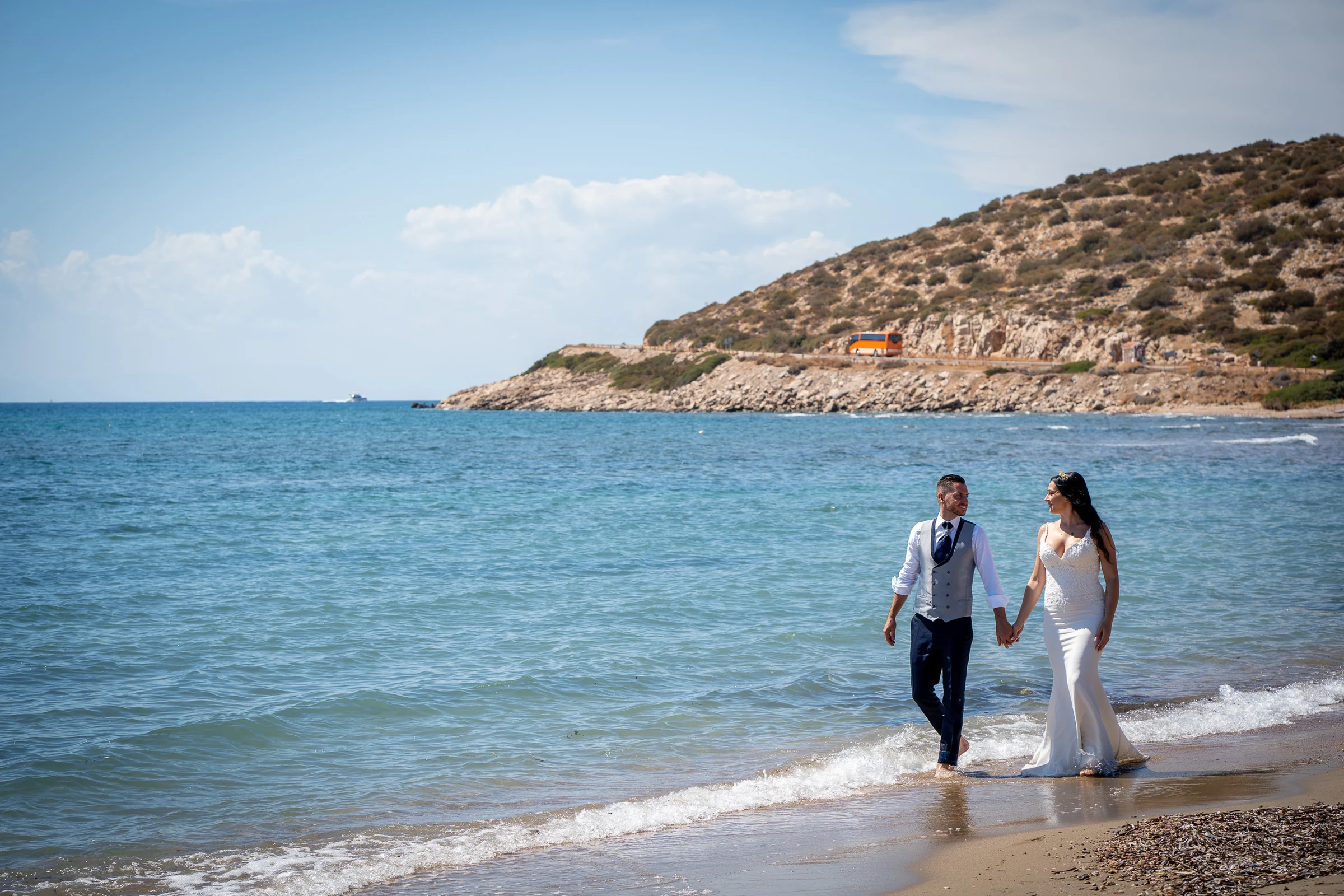 Sara y Edu pasean descalzos por la orilla de una playa griega con el mar Egeo extendiéndose a su lado durante la postboda.
