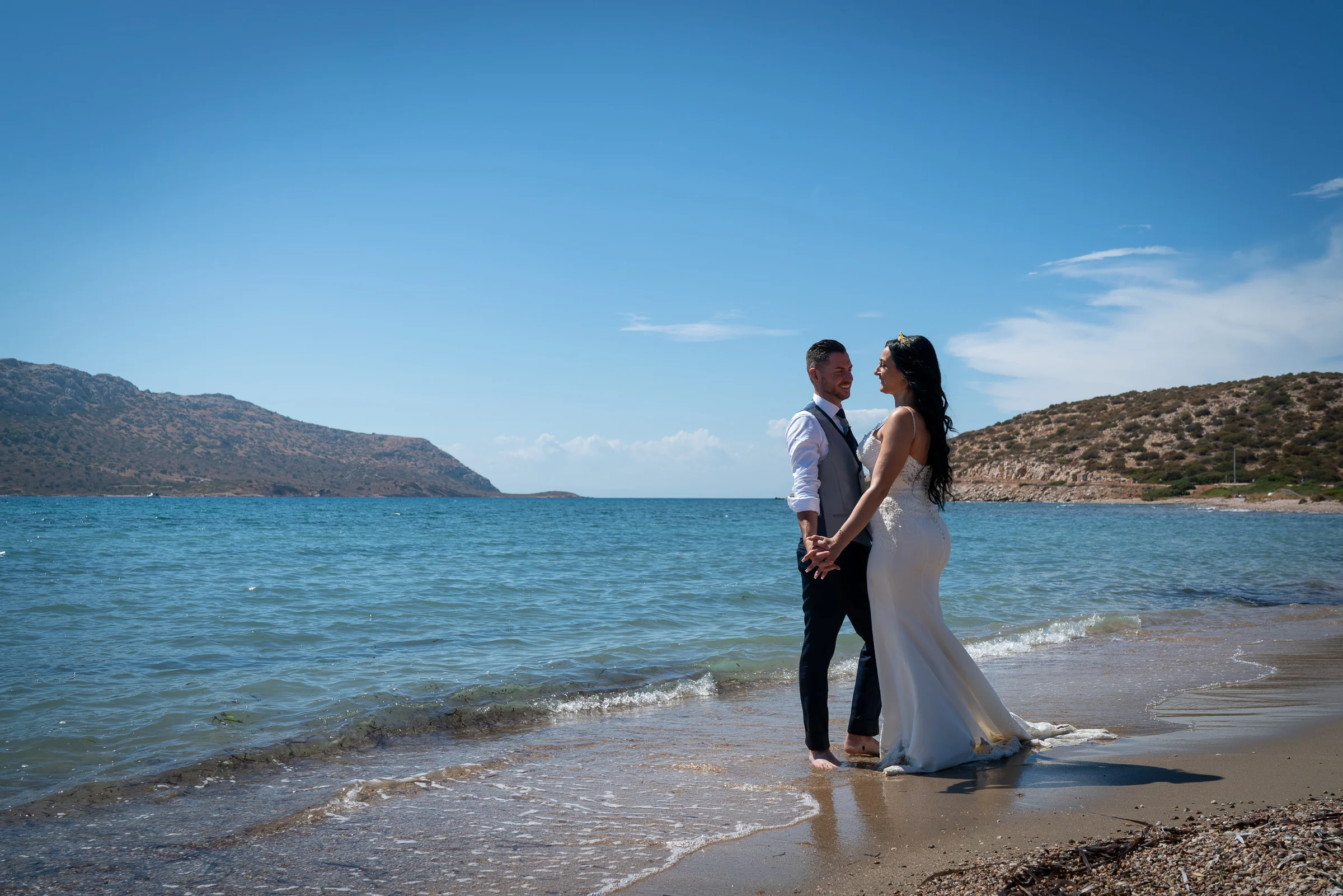 Sara y Edu se miran tomados de las manos en la orilla del mar Egeo durante su postboda en Grecia.