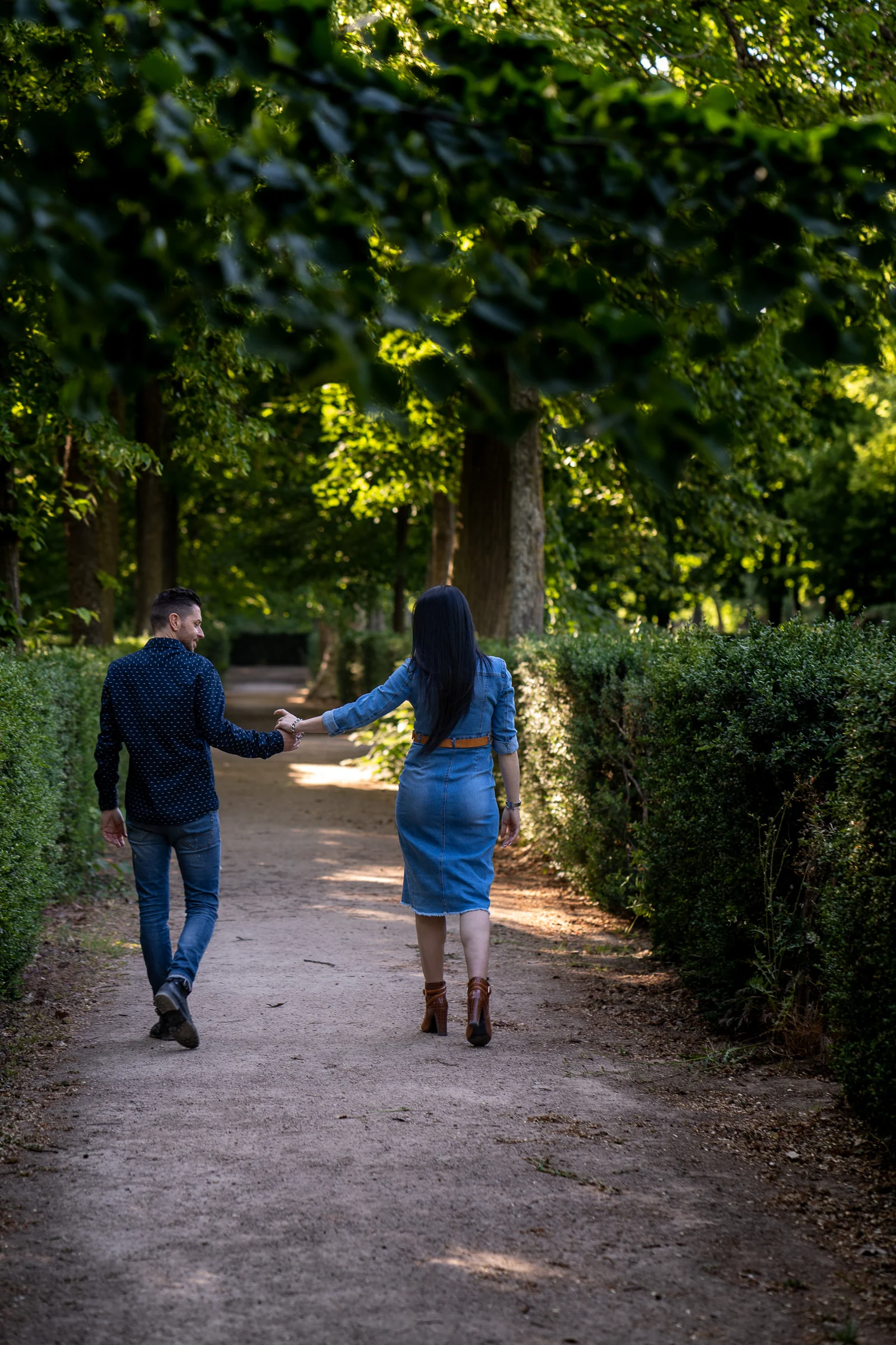 Sara y Edu se alejan caminando de la mano por un sendero entre setos durante su preboda en Aranjuez.