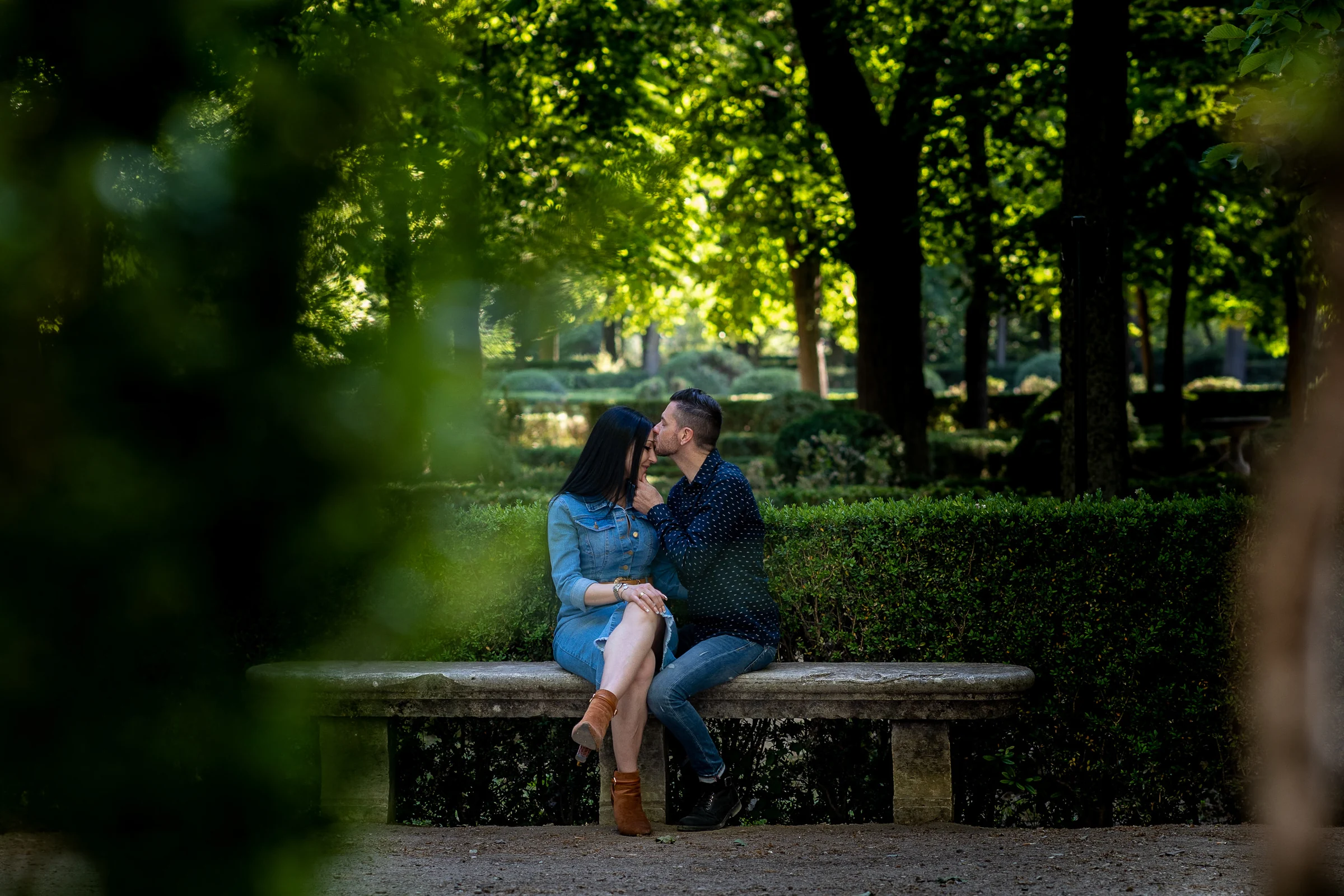Sara y Edu se besan sentados en un banco de piedra entre la vegetación de los jardines de Aranjuez.