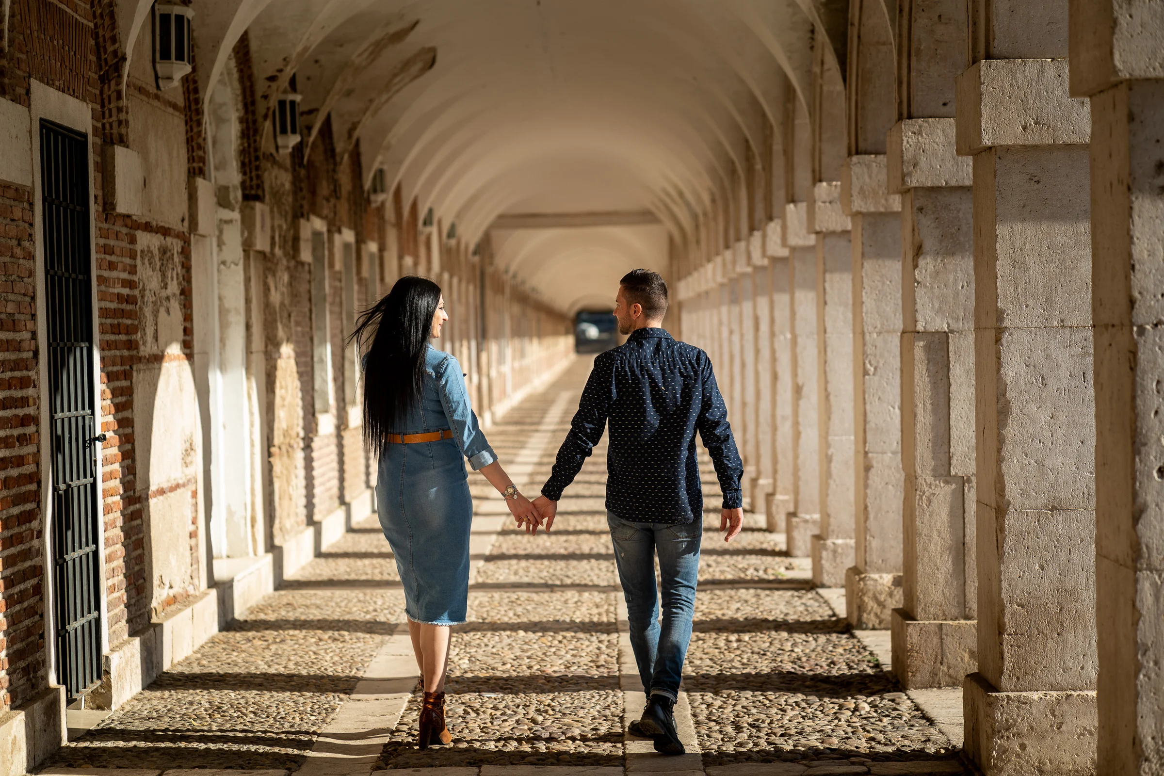 Sara y Edu caminan de la mano por una galería porticada durante su preboda en Aranjuez.