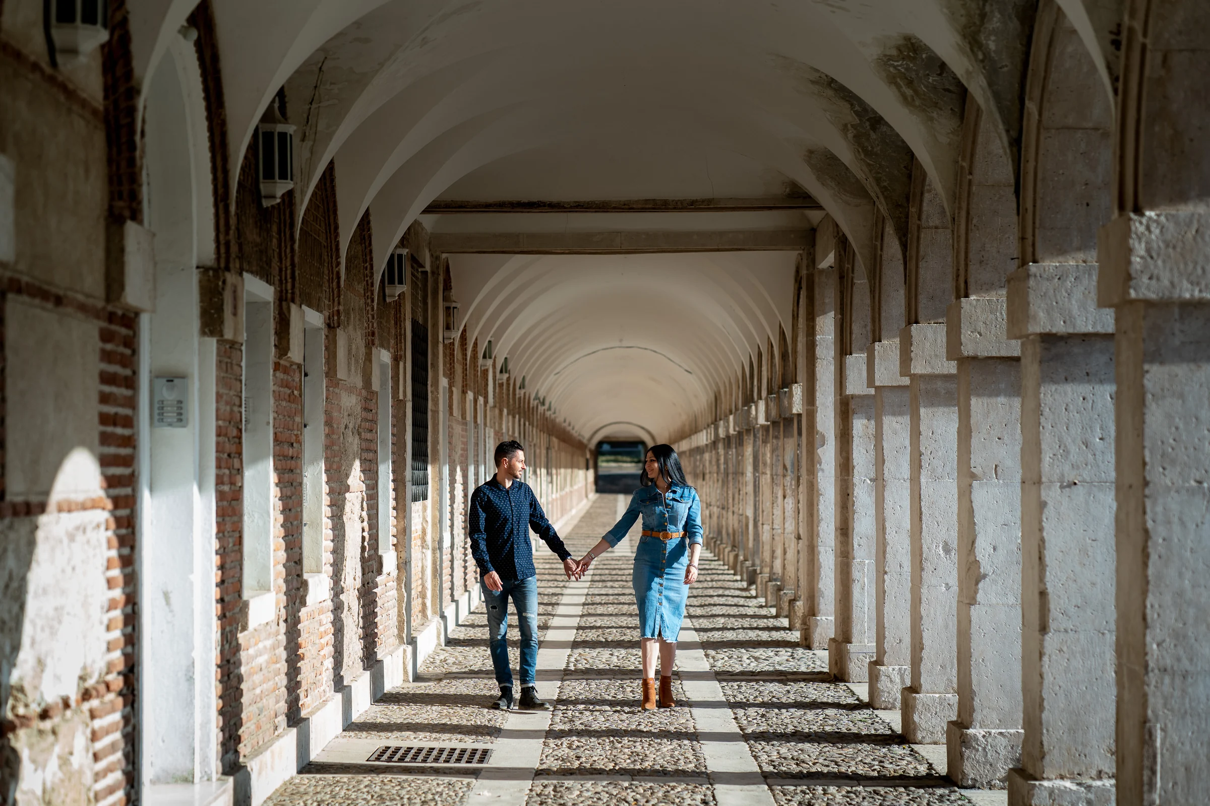 Sara y Edu caminan juntos por una galería de arcos durante su sesión de preboda en Aranjuez.