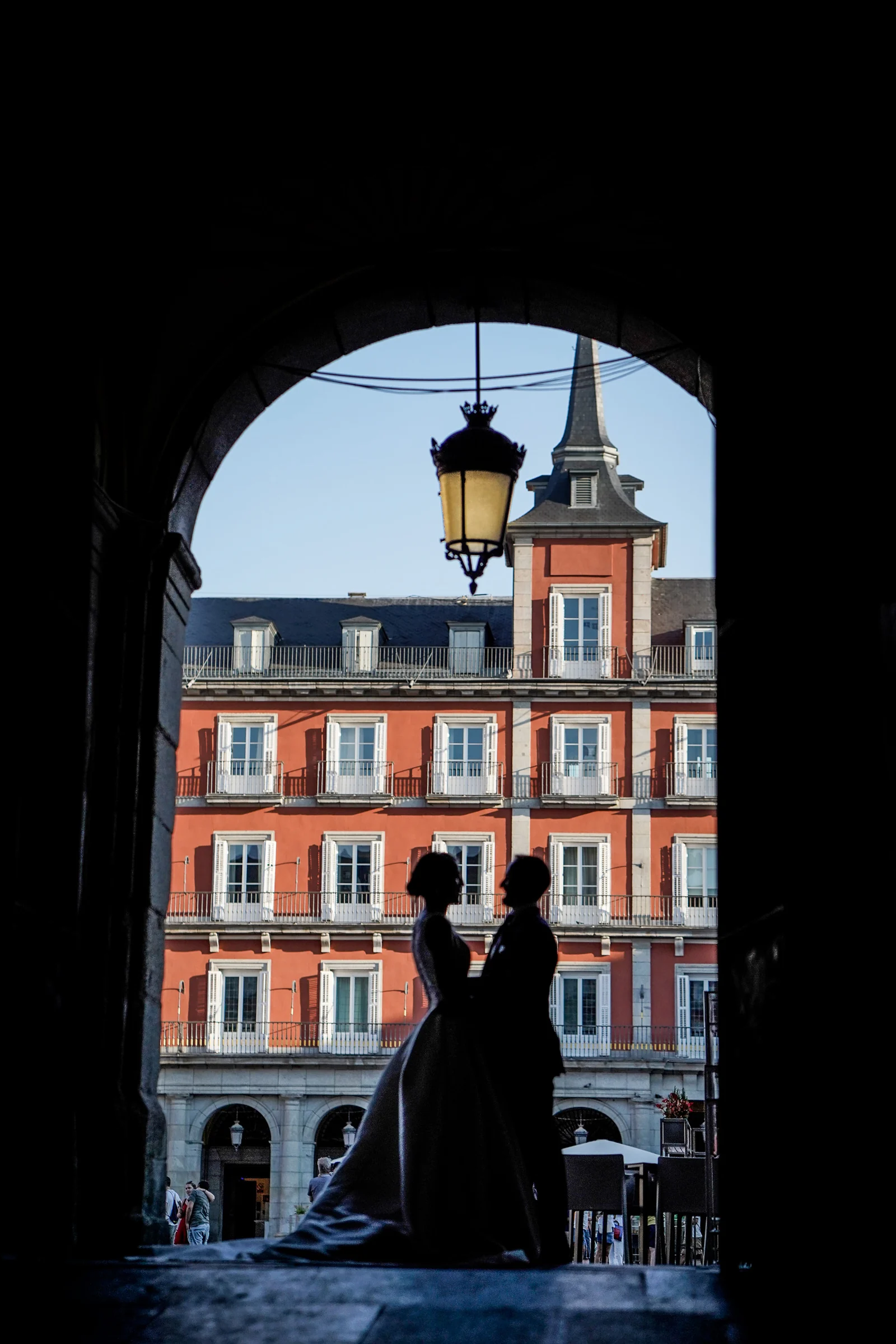 Silueta de Sara y Edu bajo uno de los arcos de la Plaza Mayor con las fachadas del centro histórico de Madrid al fondo.