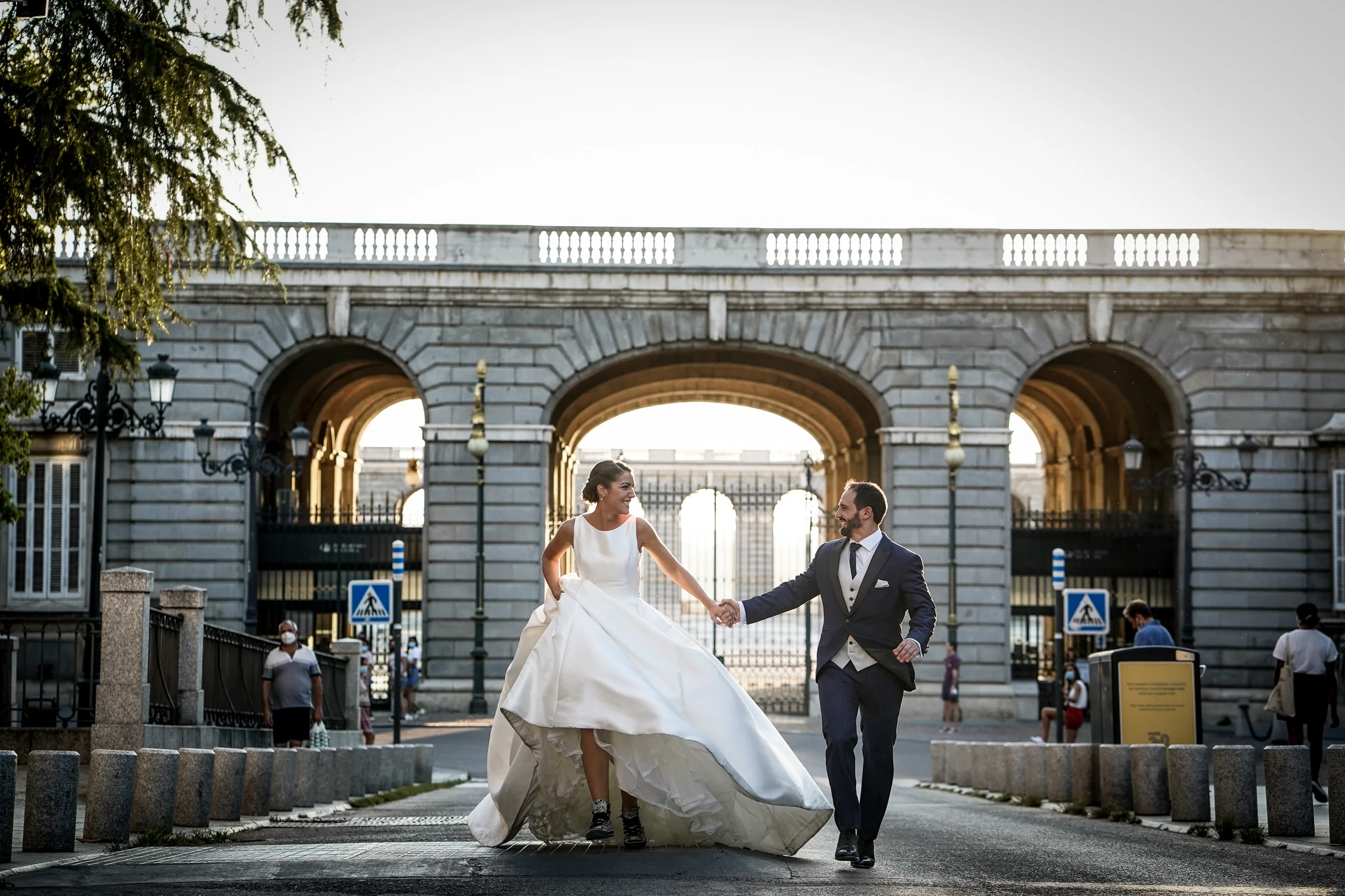 Sara y Edu caminan de la mano frente a los arcos del Palacio Real durante su sesión de postboda en Madrid.