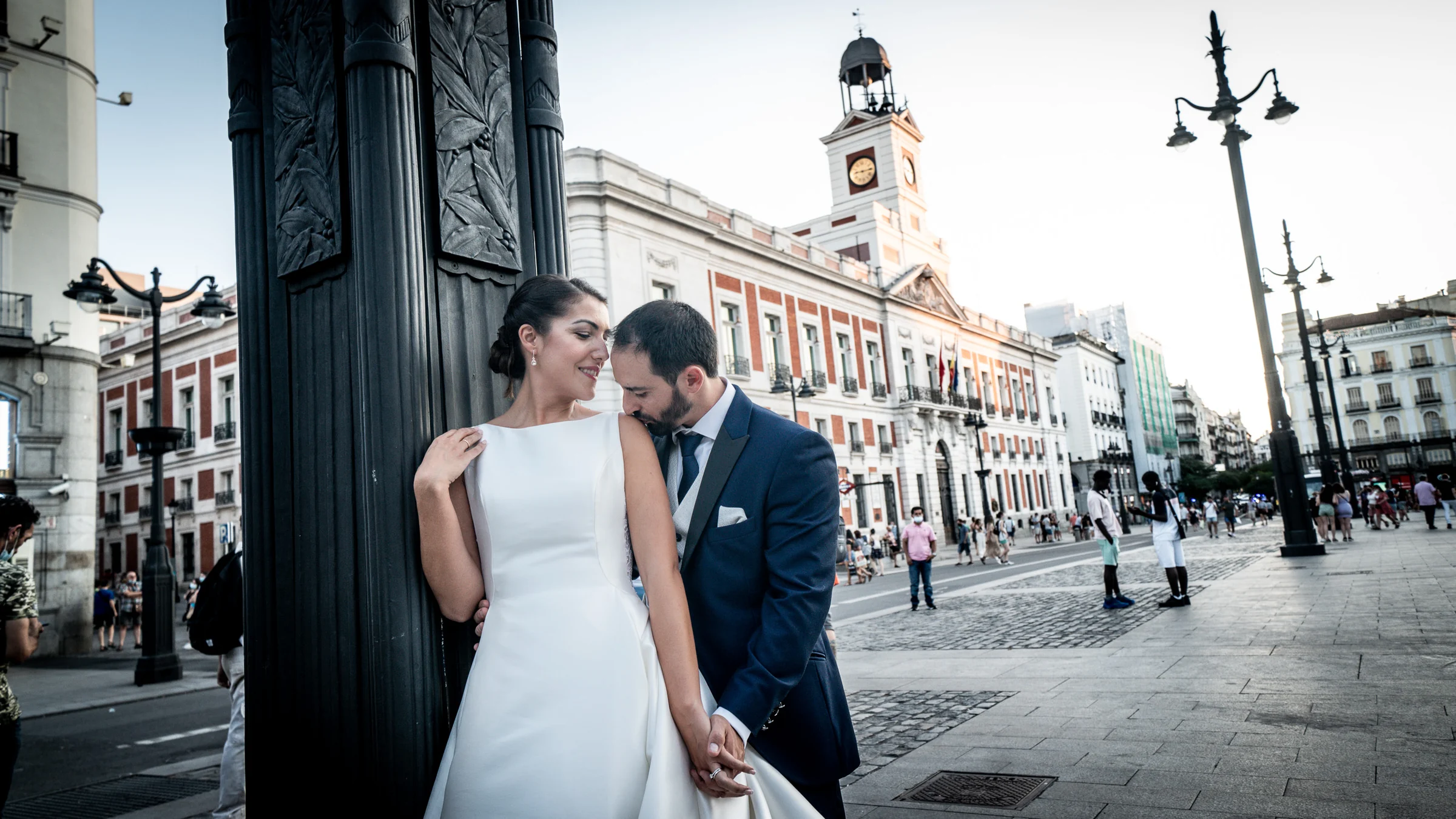 Sara y Edu se abrazan junto a una farola de la Puerta del Sol con la Real Casa de Correos al fondo durante su postboda en Madrid.