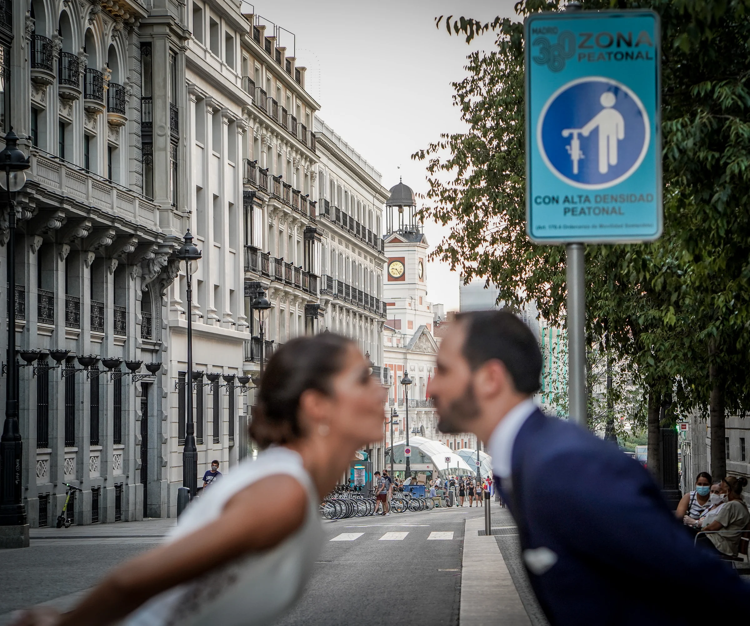Sara y Edu se acercan para besarse con la Puerta del Sol y la señal de zona peatonal al fondo durante su postboda en Madrid.