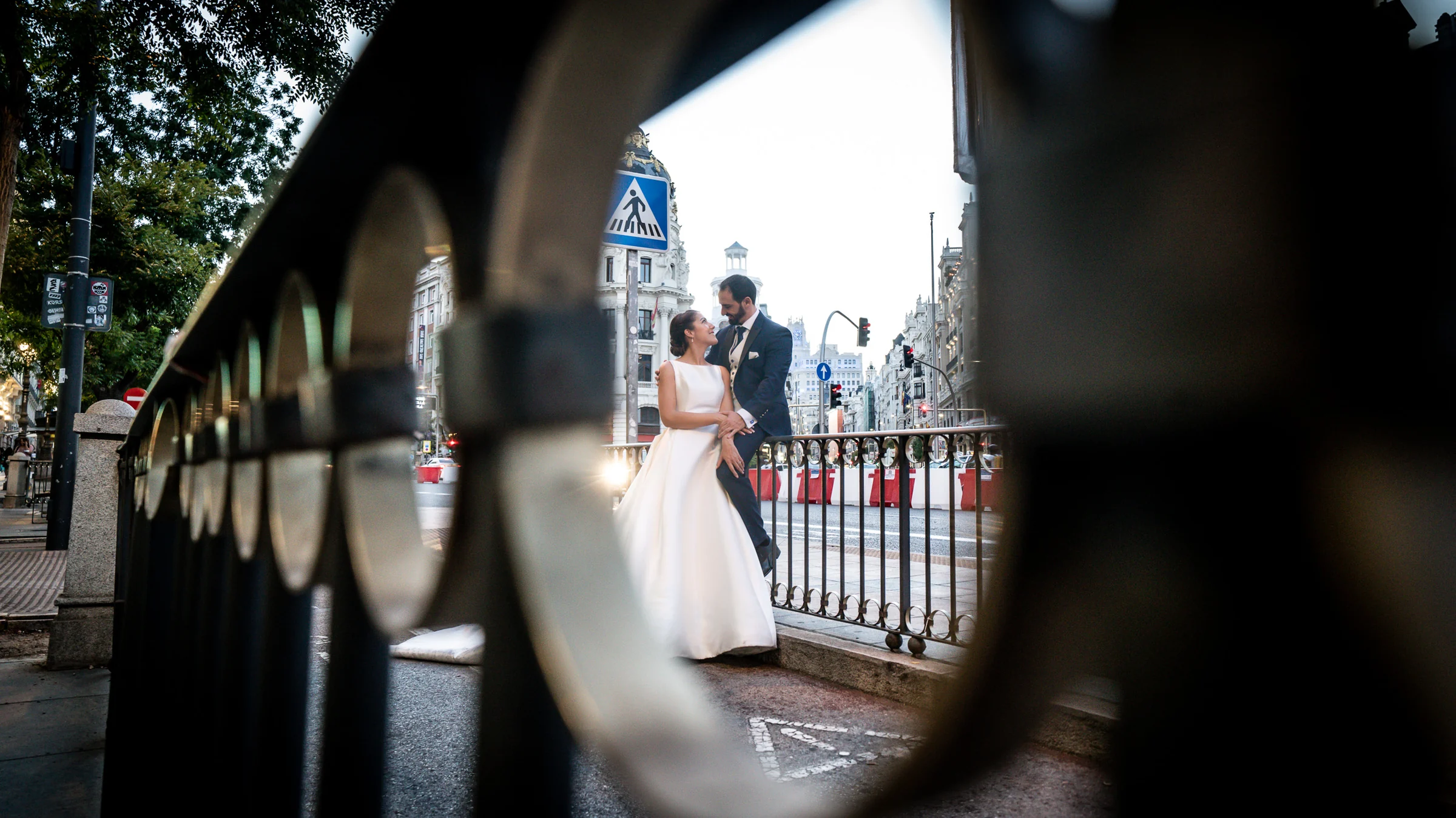 Sara y Edu aparecen enmarcados por una barandilla con el Edificio Metrópolis al fondo durante su postboda en Madrid.