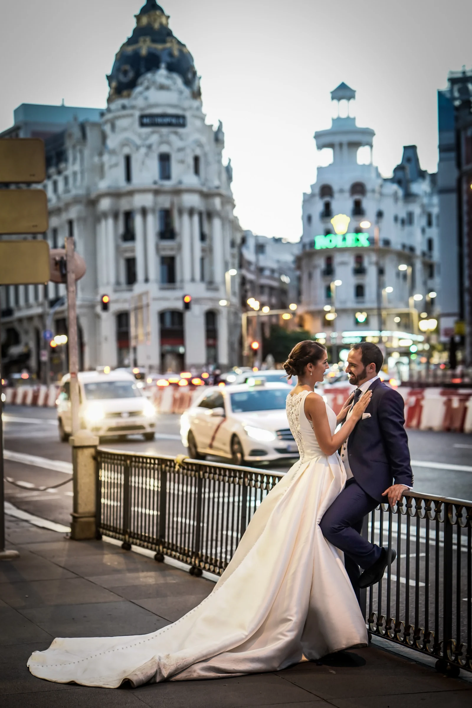 Sara y Edu posan junto a una barandilla con el Edificio Metrópolis iluminando el fondo de su postboda en Madrid.