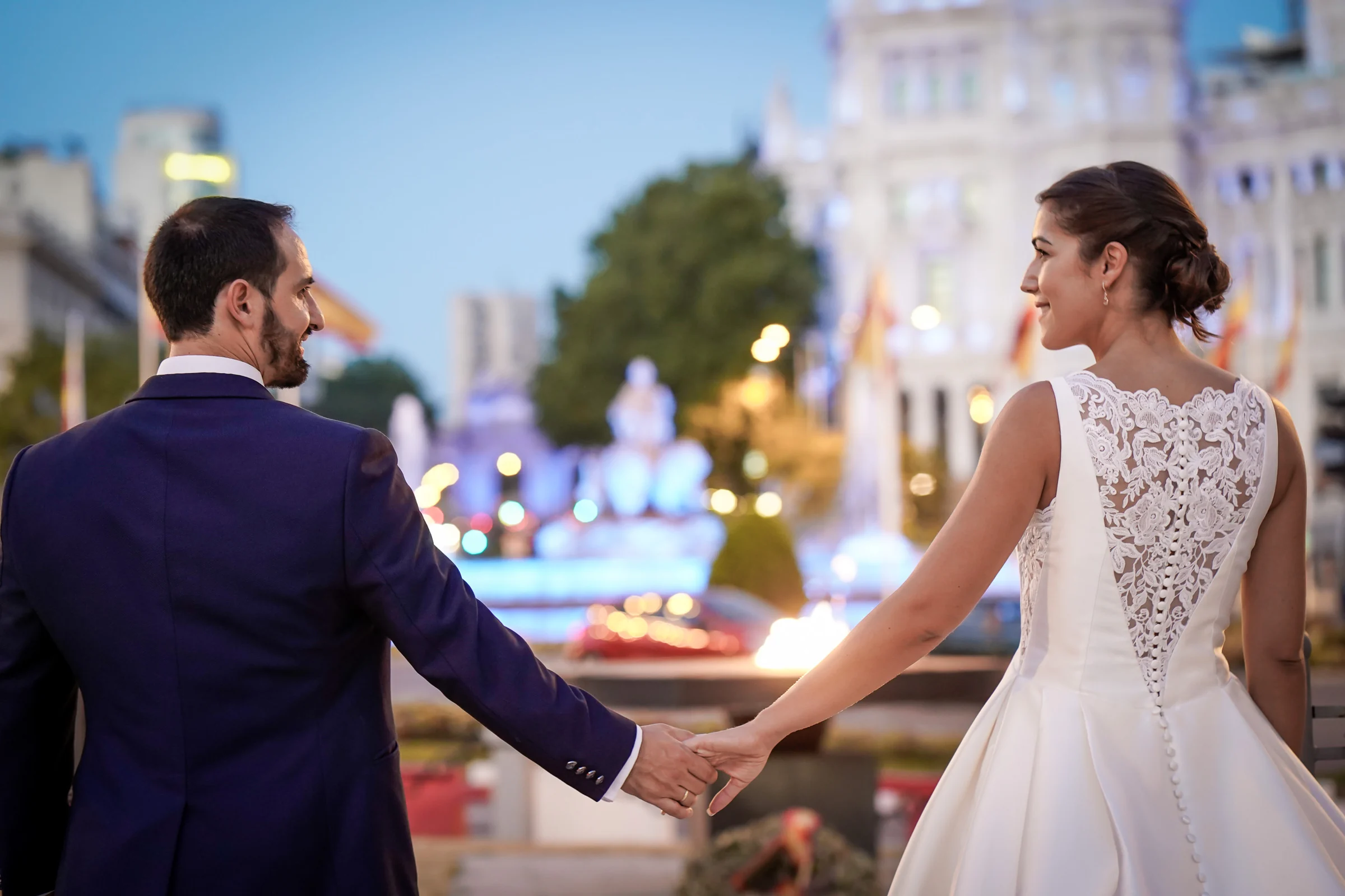 Sara y Edu se dan la mano frente a las luces de la fuente de Cibeles durante su postboda al anochecer en Madrid.