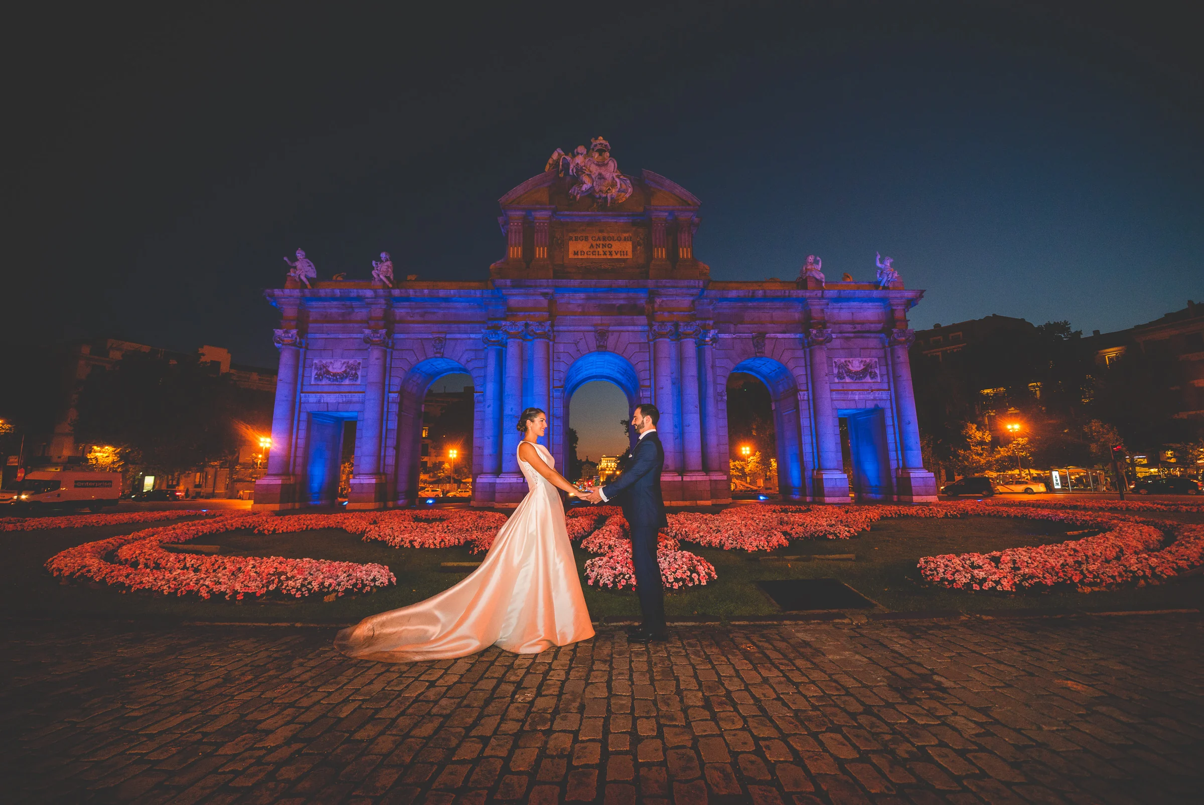 Sara y Edu se miran frente a la Puerta de Alcalá iluminada de azul durante su postboda nocturna en Madrid.