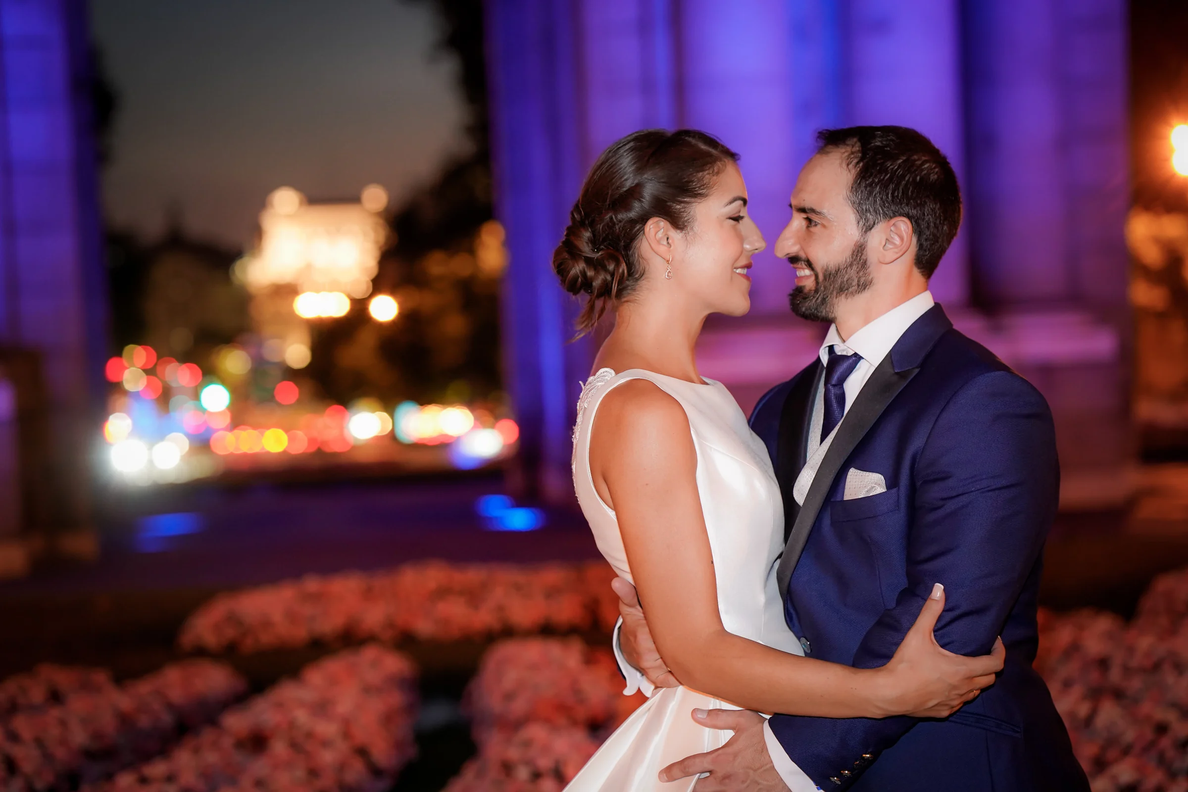 Sara y Edu se abrazan con la Puerta de Alcalá iluminada al fondo durante su postboda nocturna en Madrid.