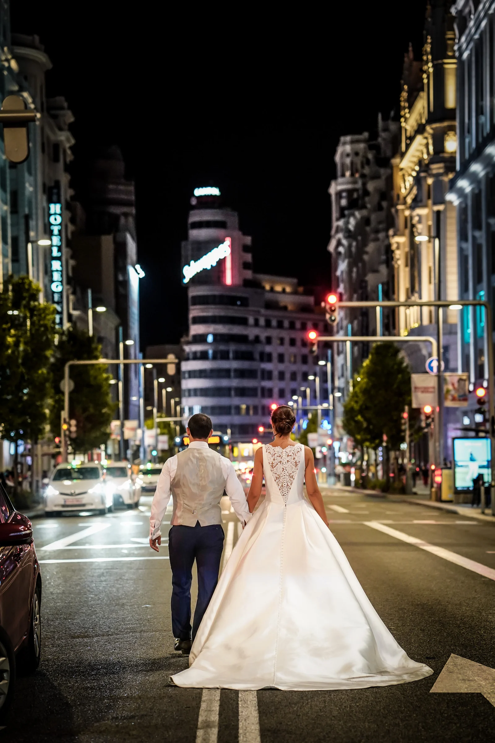 Sara y Edu caminan de espaldas por la Gran Vía iluminada durante su postboda nocturna en Madrid.