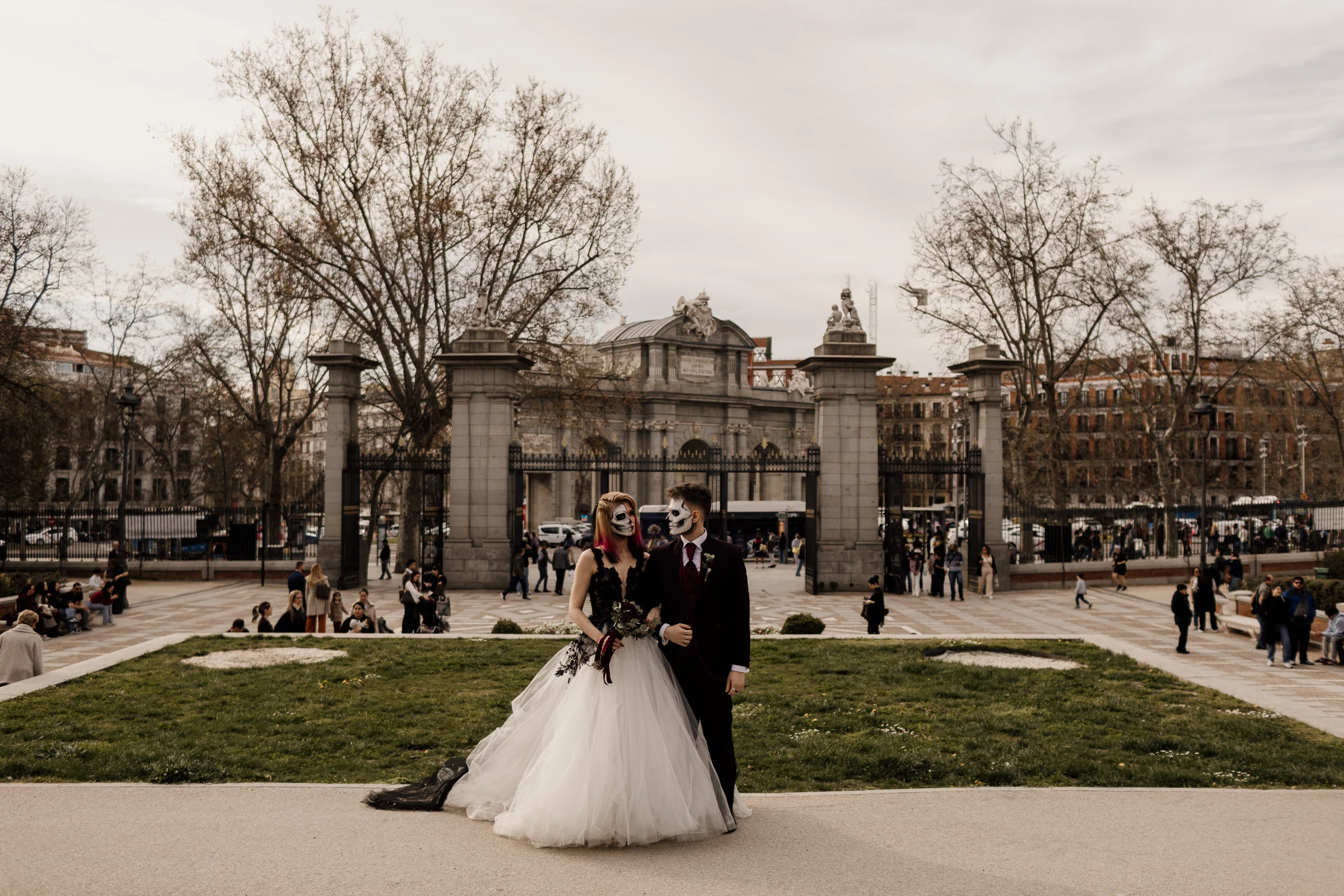 Silvia y Rubén posan con maquillaje de calavera frente a una entrada histórica del Parque de El Retiro durante su postboda gótica en Madrid.