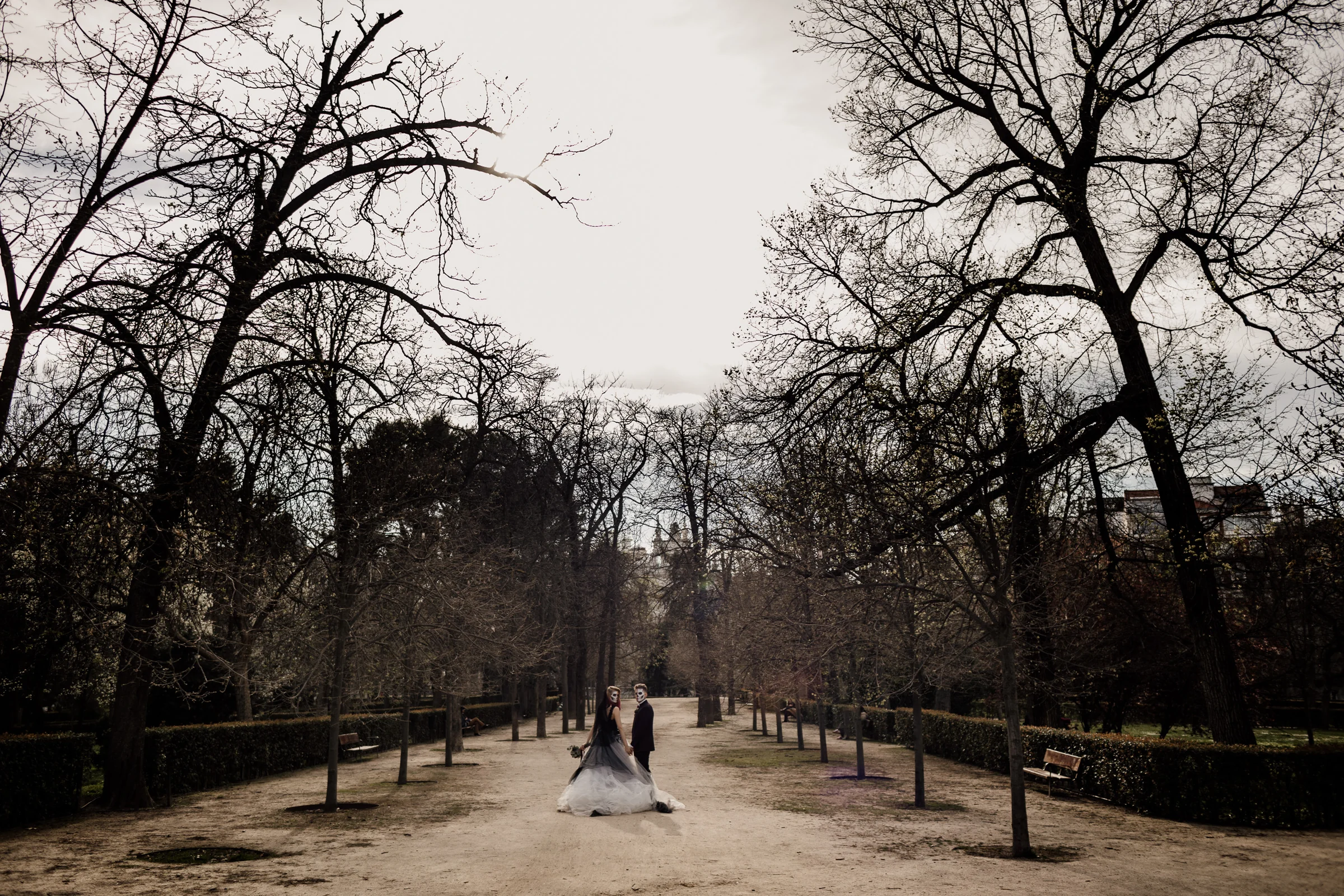 Silvia y Rubén aparecen a distancia en un paseo arbolado del Parque de El Retiro durante su postboda gótica en Madrid.