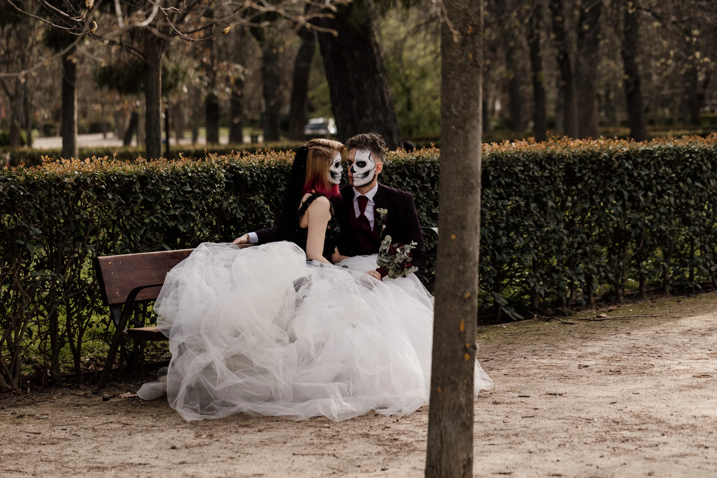 Silvia y Rubén se miran sentados en un banco entre setos y árboles durante su postboda gótica en El Retiro.