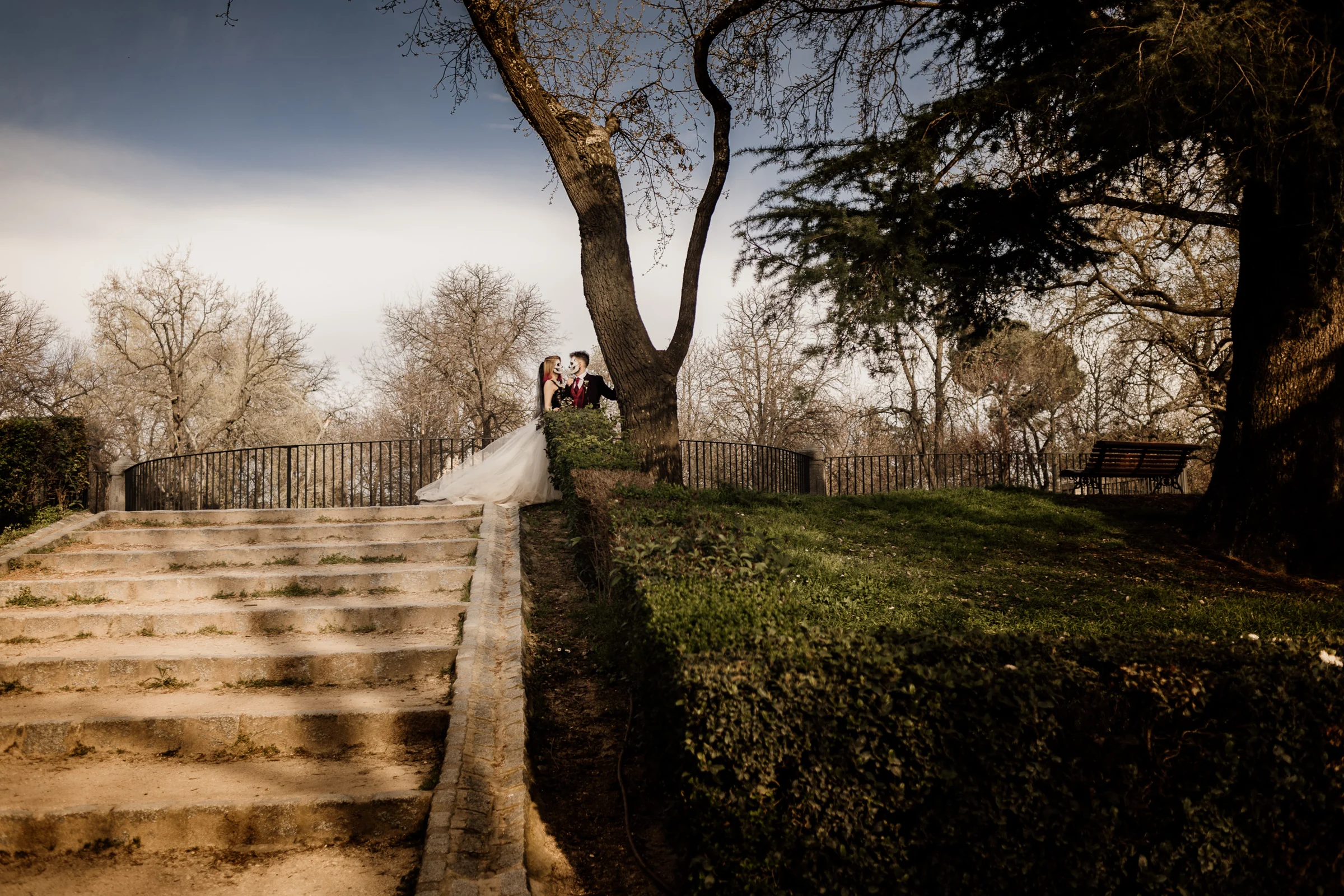 Silvia y Rubén se abrazan junto a un gran árbol sobre una escalinata del Parque de El Retiro durante su postboda gótica.