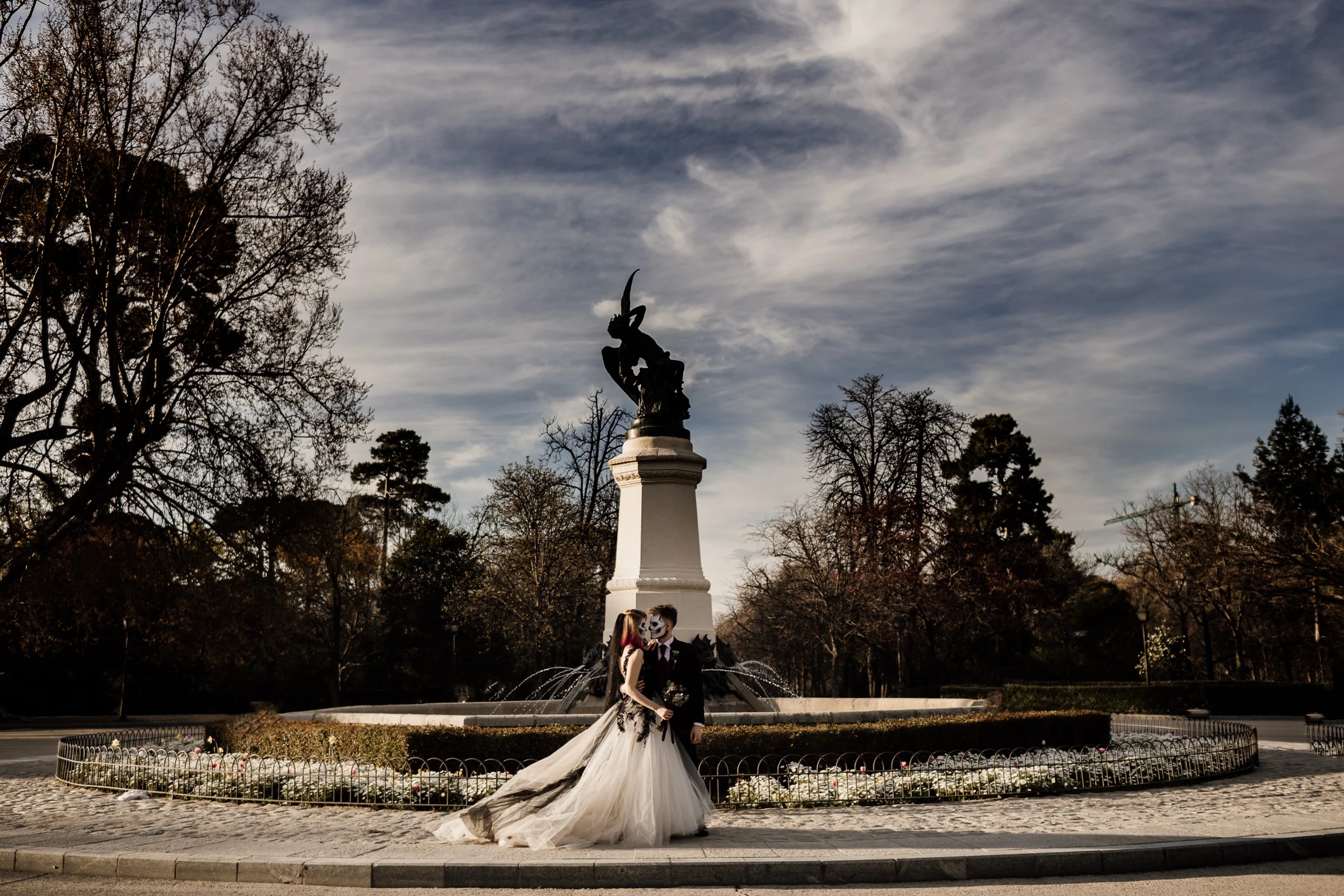 Silvia y Rubén posan delante de la Fuente del Ángel Caído en el Parque de El Retiro durante su postboda gótica en Madrid.