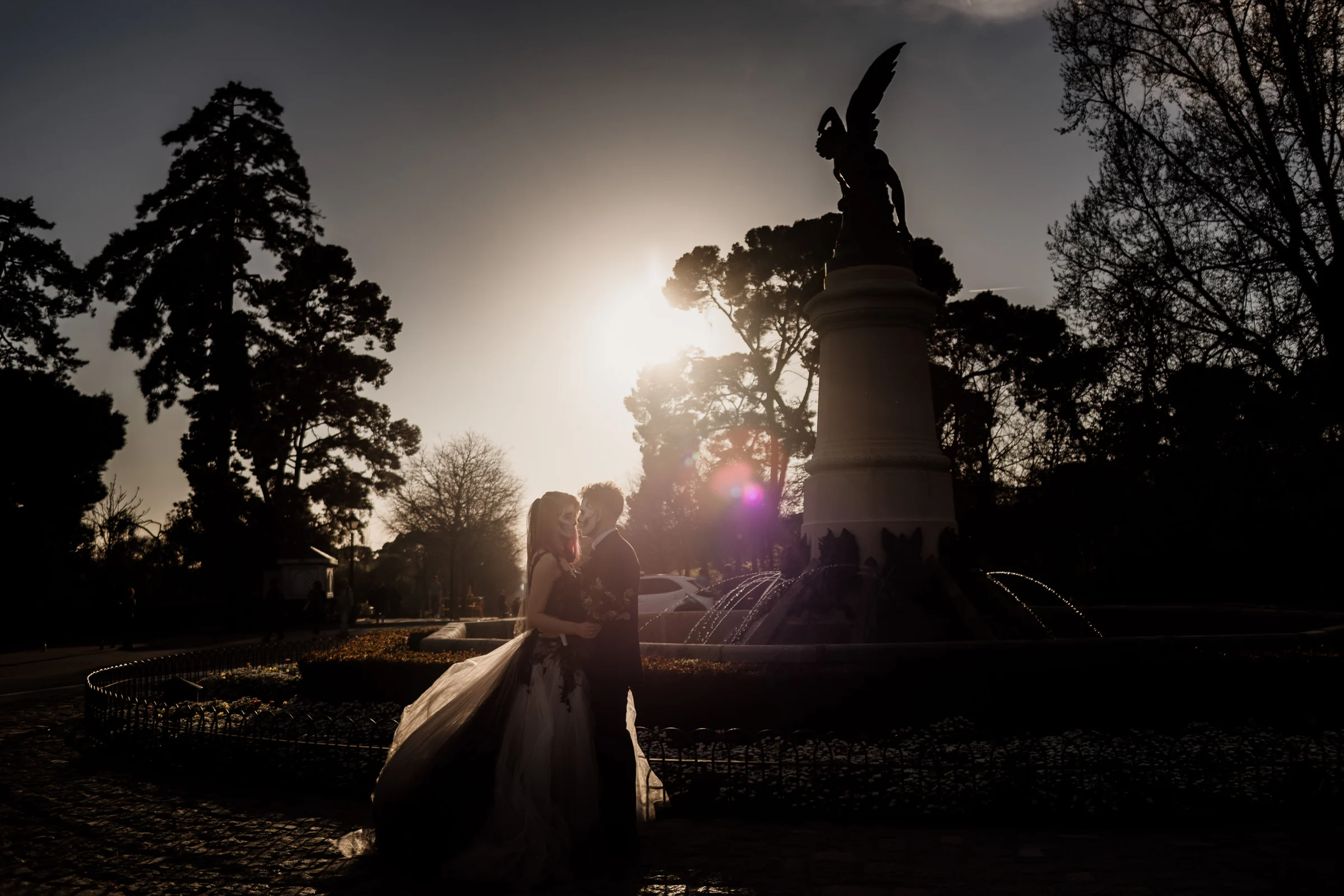 Silvia y Rubén aparecen a contraluz junto a la Fuente del Ángel Caído con el sol bajo detrás de la pareja en El Retiro.