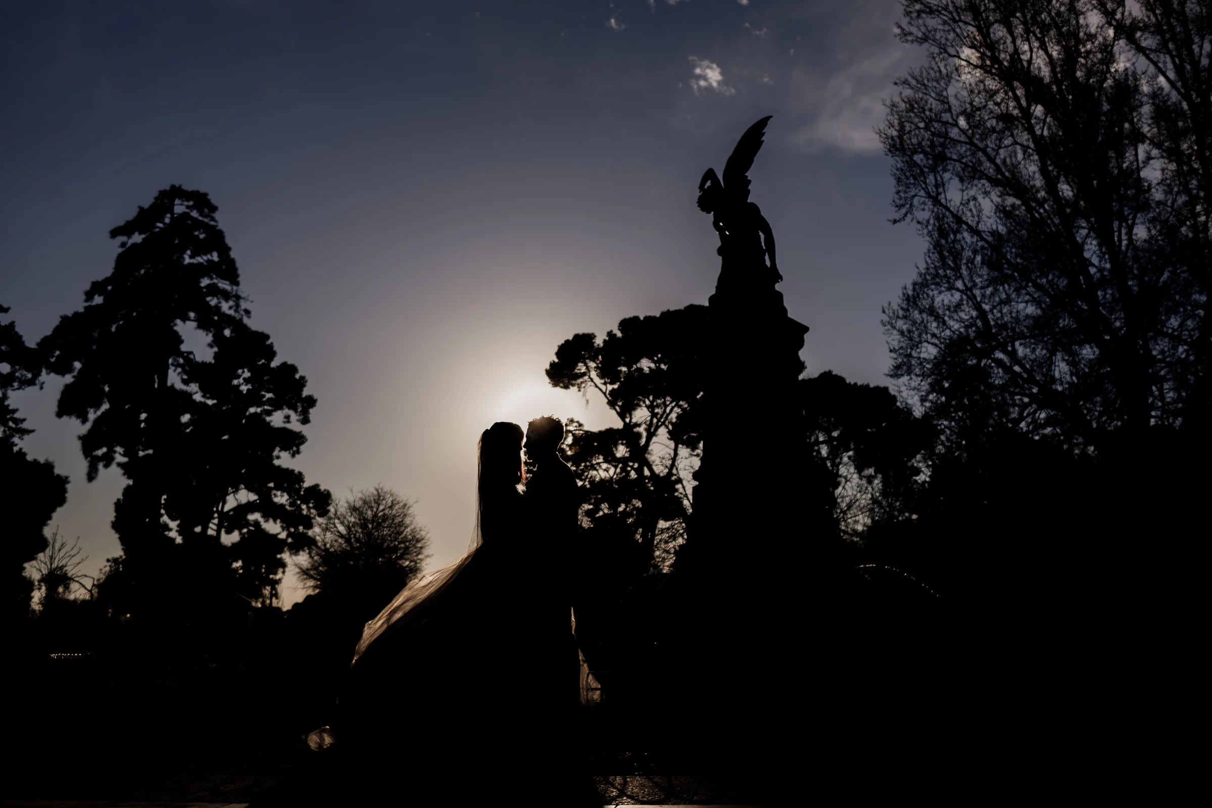 Silvia y Rubén se besan en silueta frente a la Fuente del Ángel Caído al caer la luz en el Parque de El Retiro.